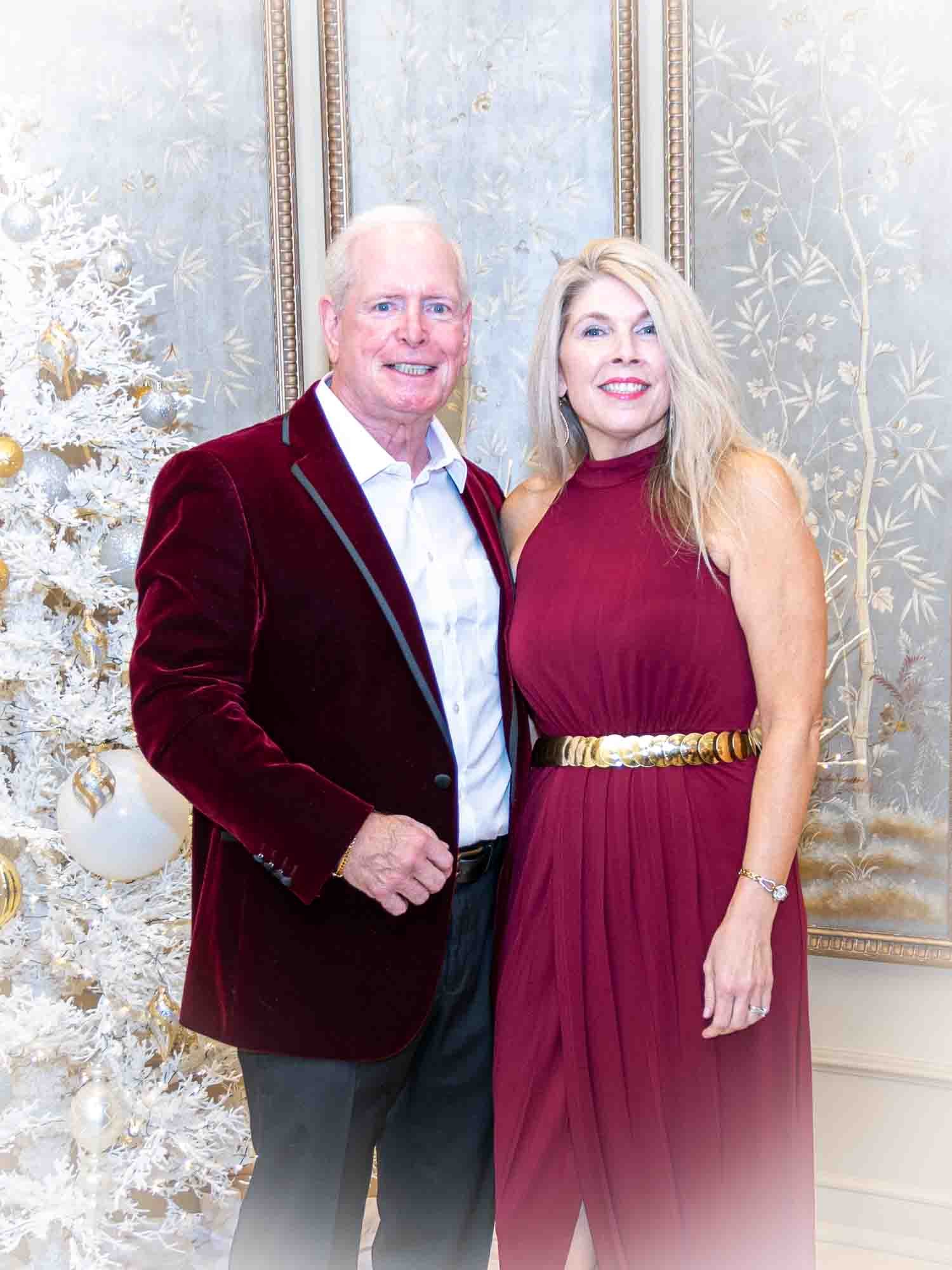 A couple posing together indoors, dressed in formal attire with a decorated white Christmas tree in the background. The man is wearing a burgundy velvet jacket, while the woman is in a burgundy dress.