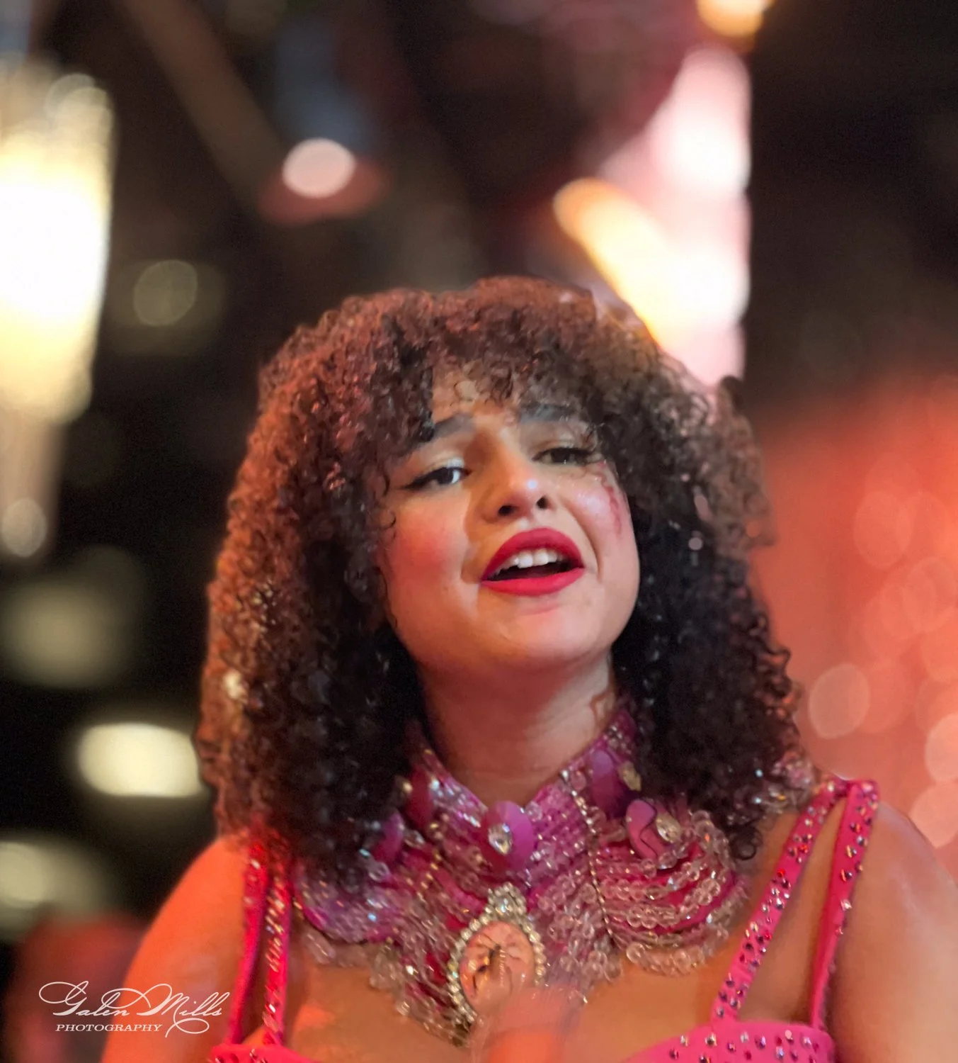 Person performing in colorful, embellished outfit with curly hair and red lipstick in a dimly lit setting.
