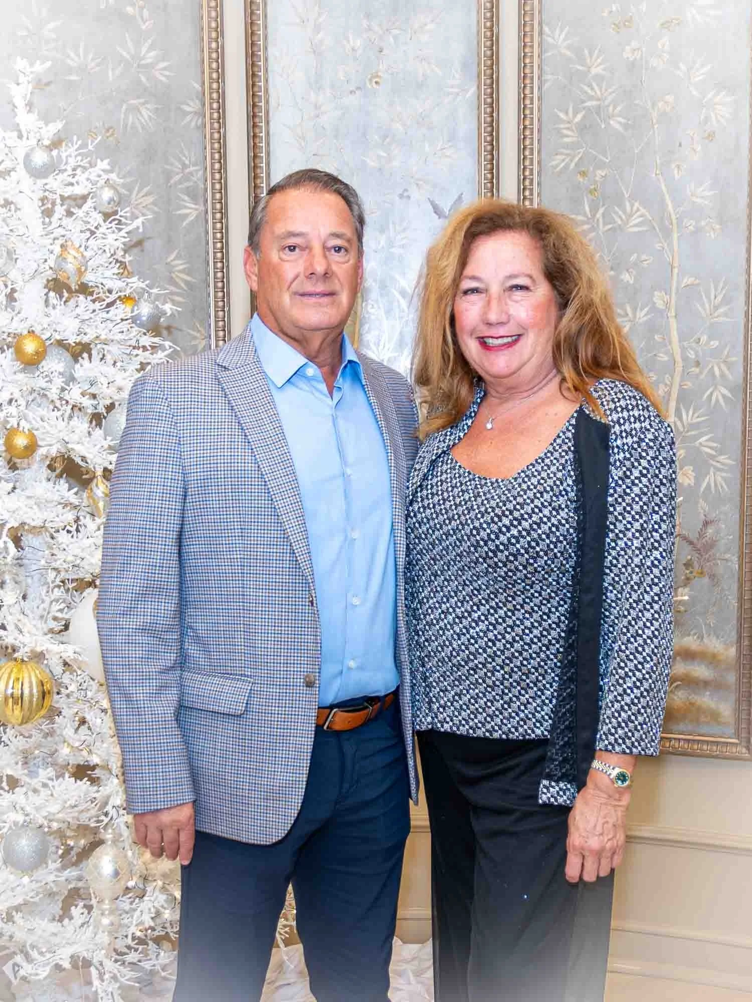 A man and woman standing together indoors, wearing formal attire. They are next to a decorated white Christmas tree. The background features elegant wallpaper with nature patterns.