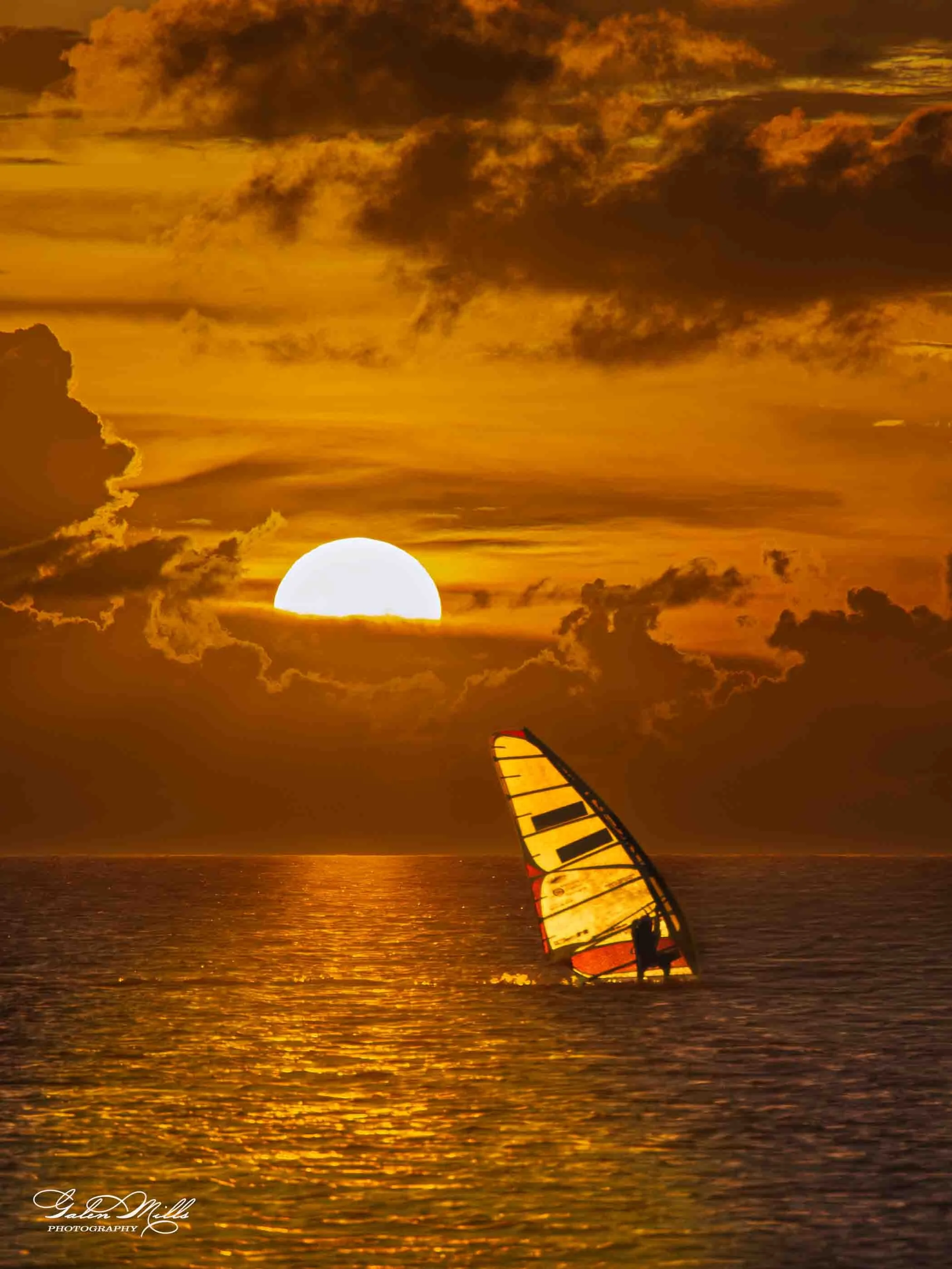A person windsurfing on the ocean during sunset with a bright sun and a cloudy sky.