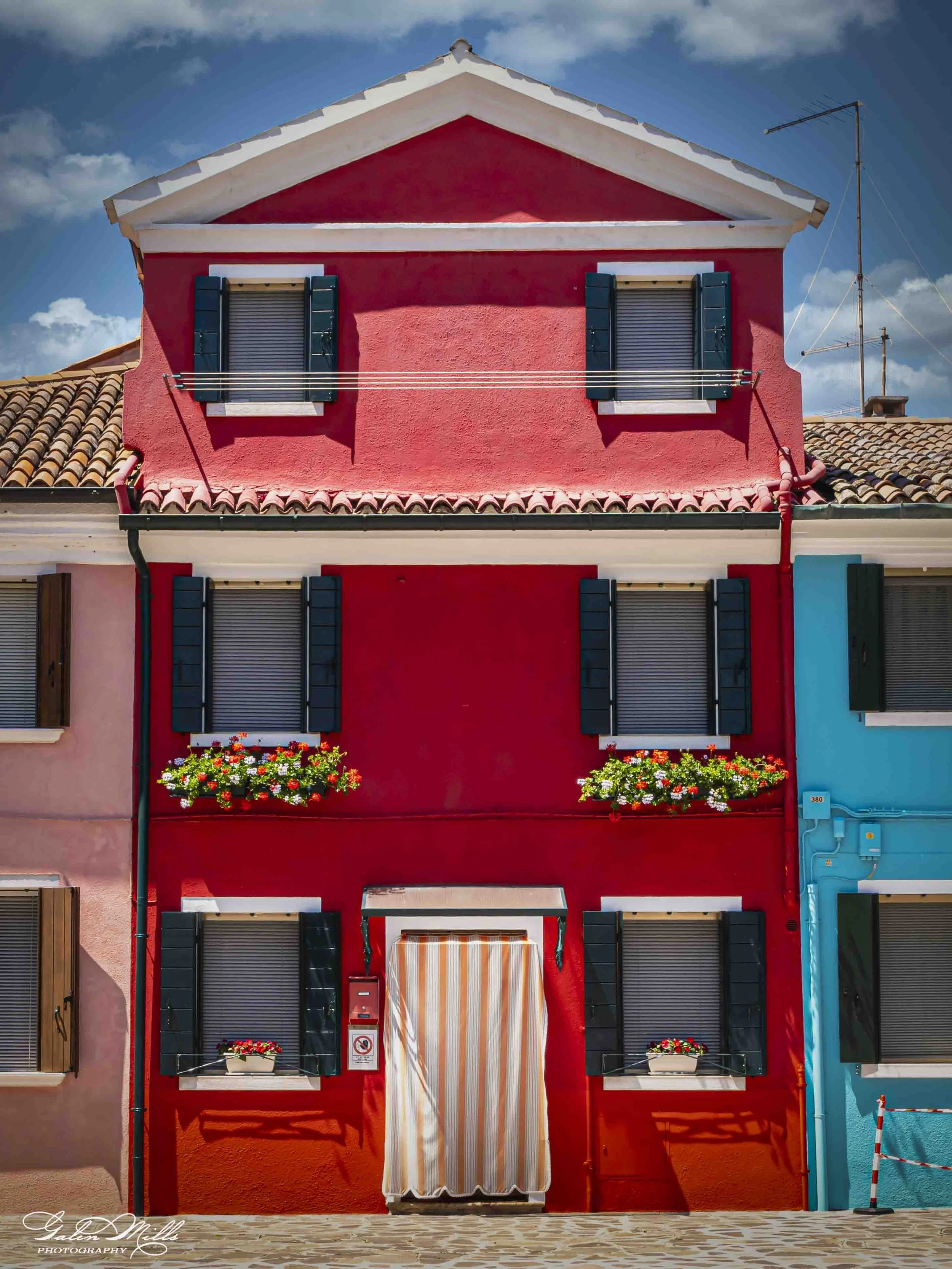 Colorful building with a pink and turquoise facade, black window shutters, flower boxes, and a striped awning, in a vibrant seaside town.