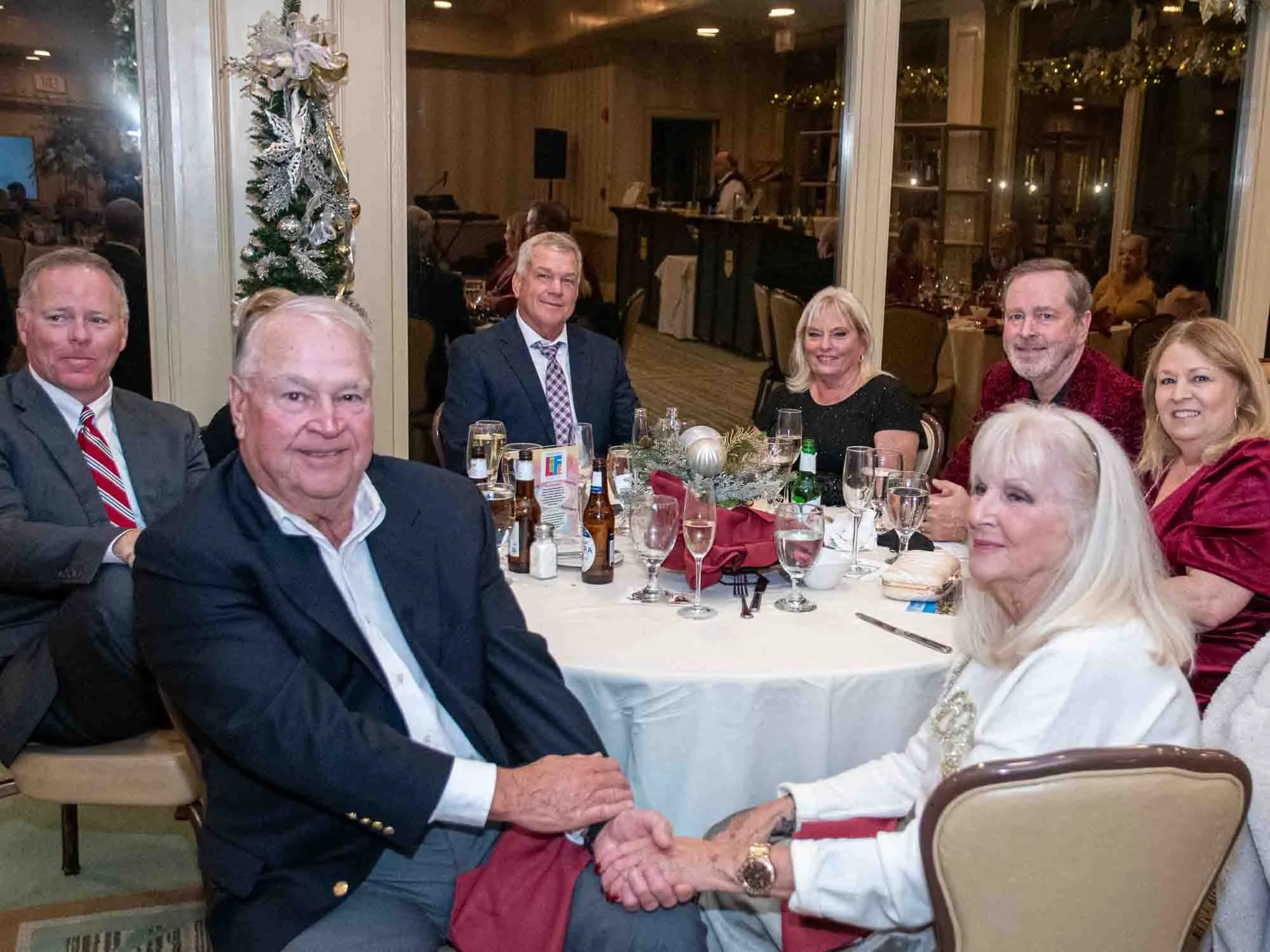Group of people sitting at a round table during a formal event in a decorated room with a small Christmas tree in the background.
