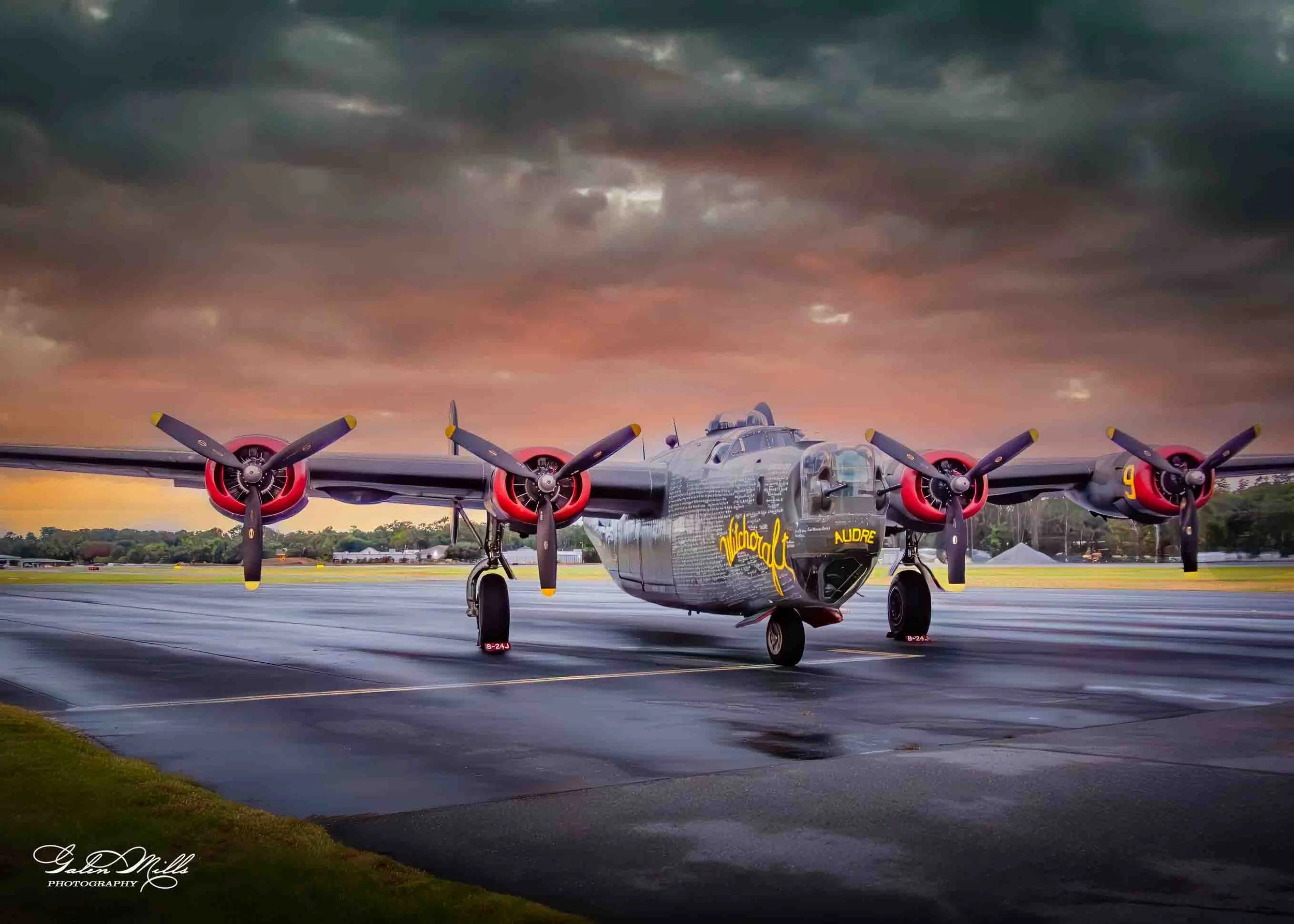 A military aircraft with four propellers on a wet runway at sunset, with dark clouds overhead.