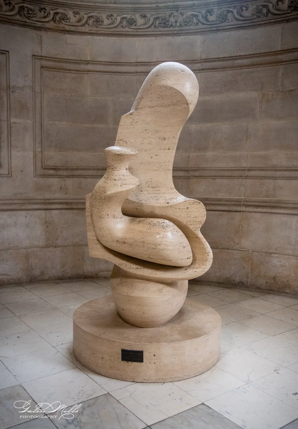 Abstract stone sculpture with smooth curves and spherical elements mounted on a circular base in a decorative interior setting.