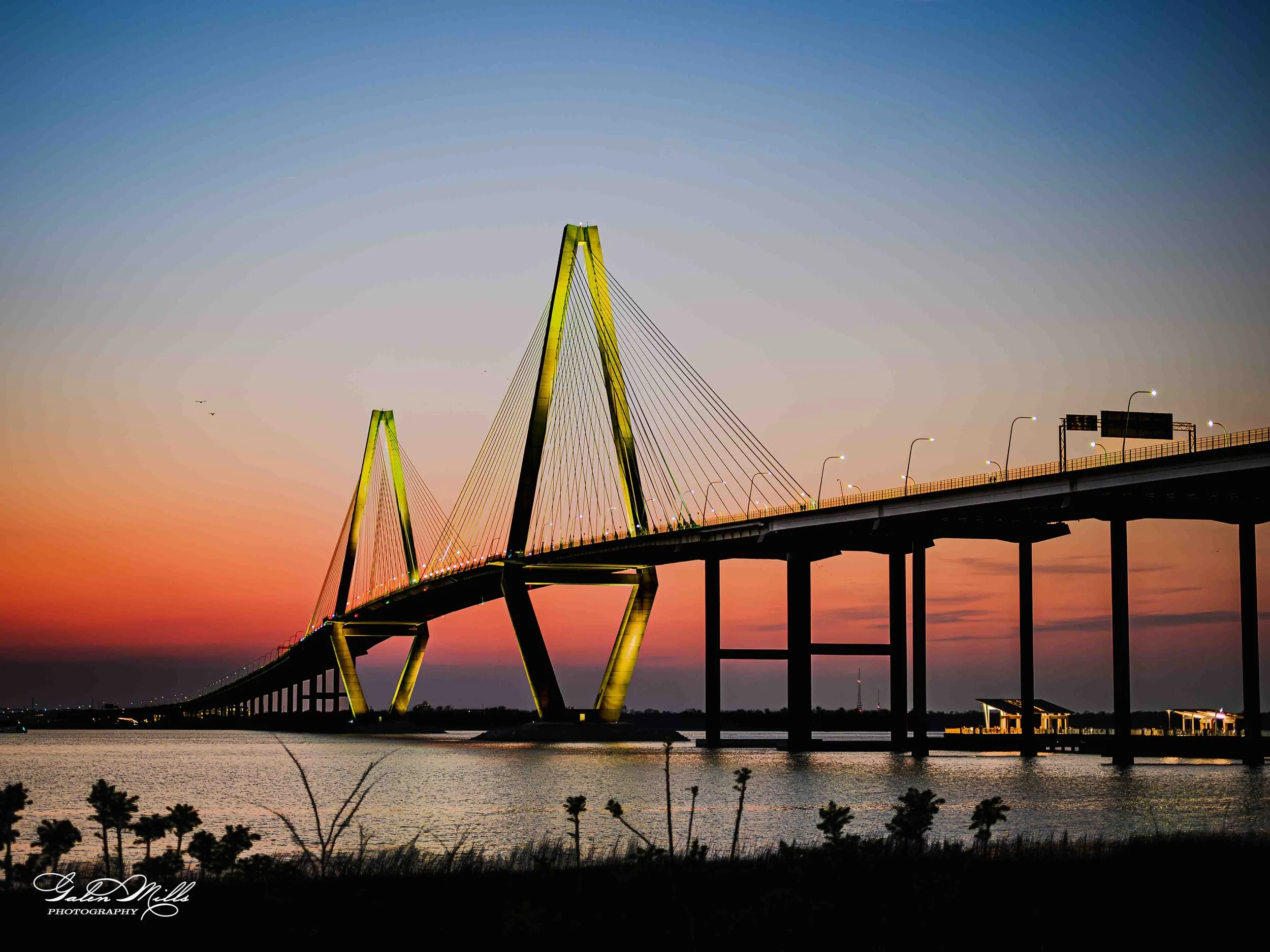 A bridge illuminated at sunset over a body of water, with a colorful sky and silhouetted vegetation in the foreground.