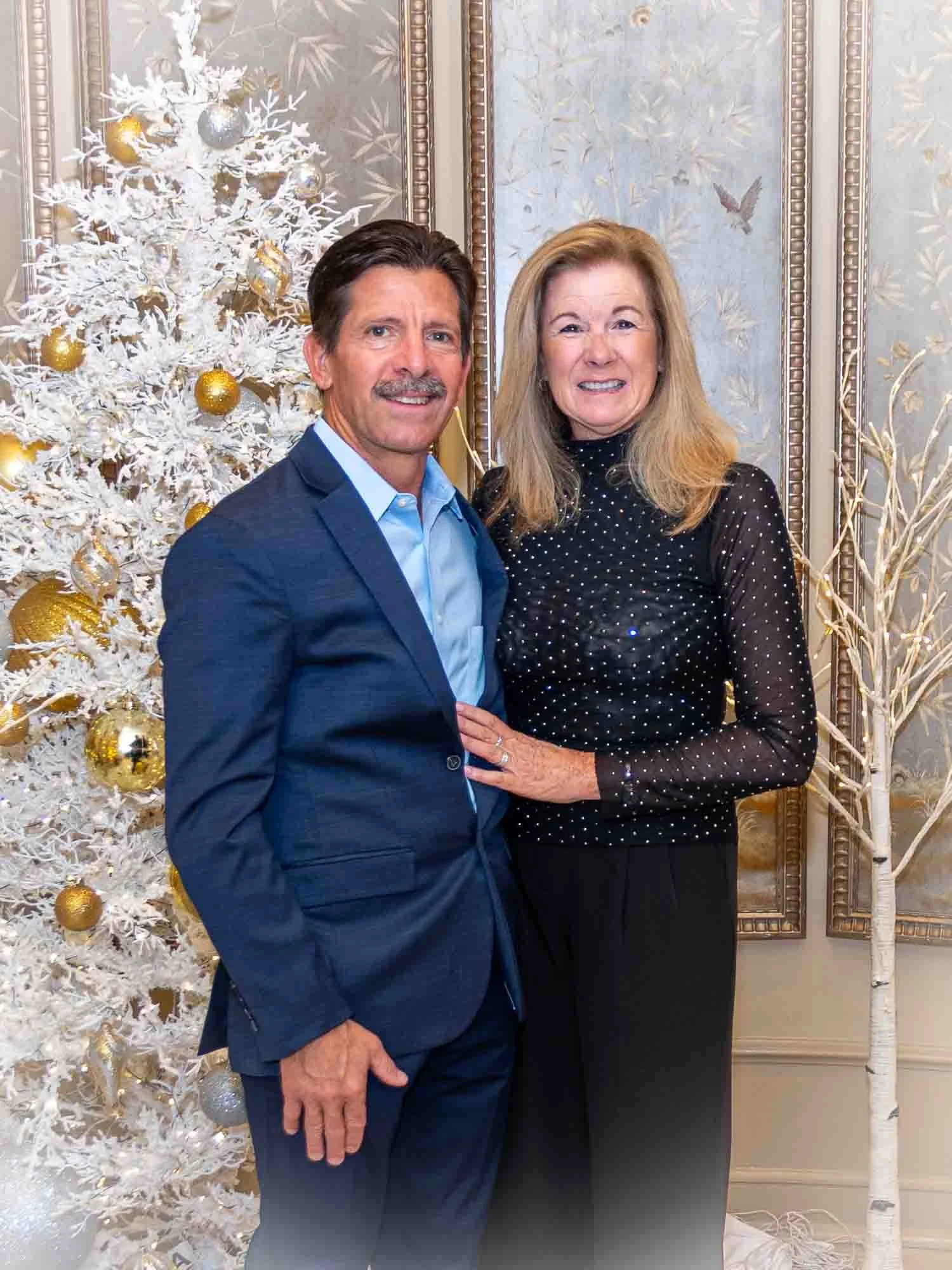 A couple posing in front of a white Christmas tree decorated with gold and silver ornaments.