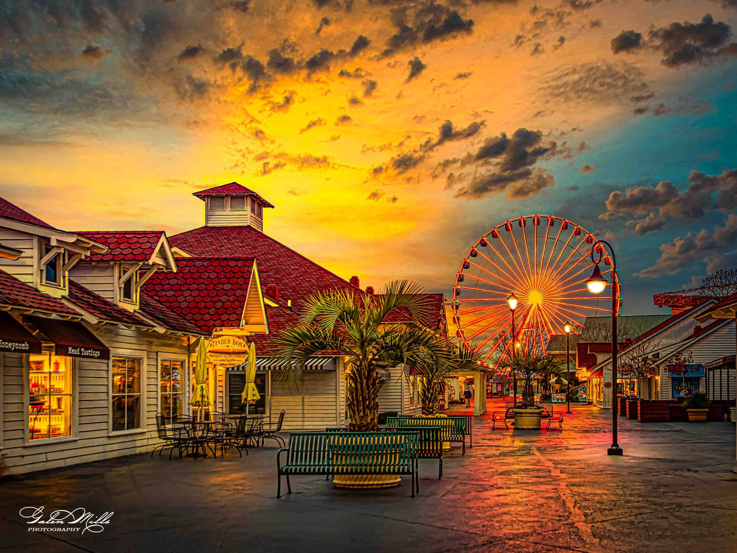 An empty outdoor shopping area at sunset with palm trees, benches, shops, and a large Ferris wheel in the background.