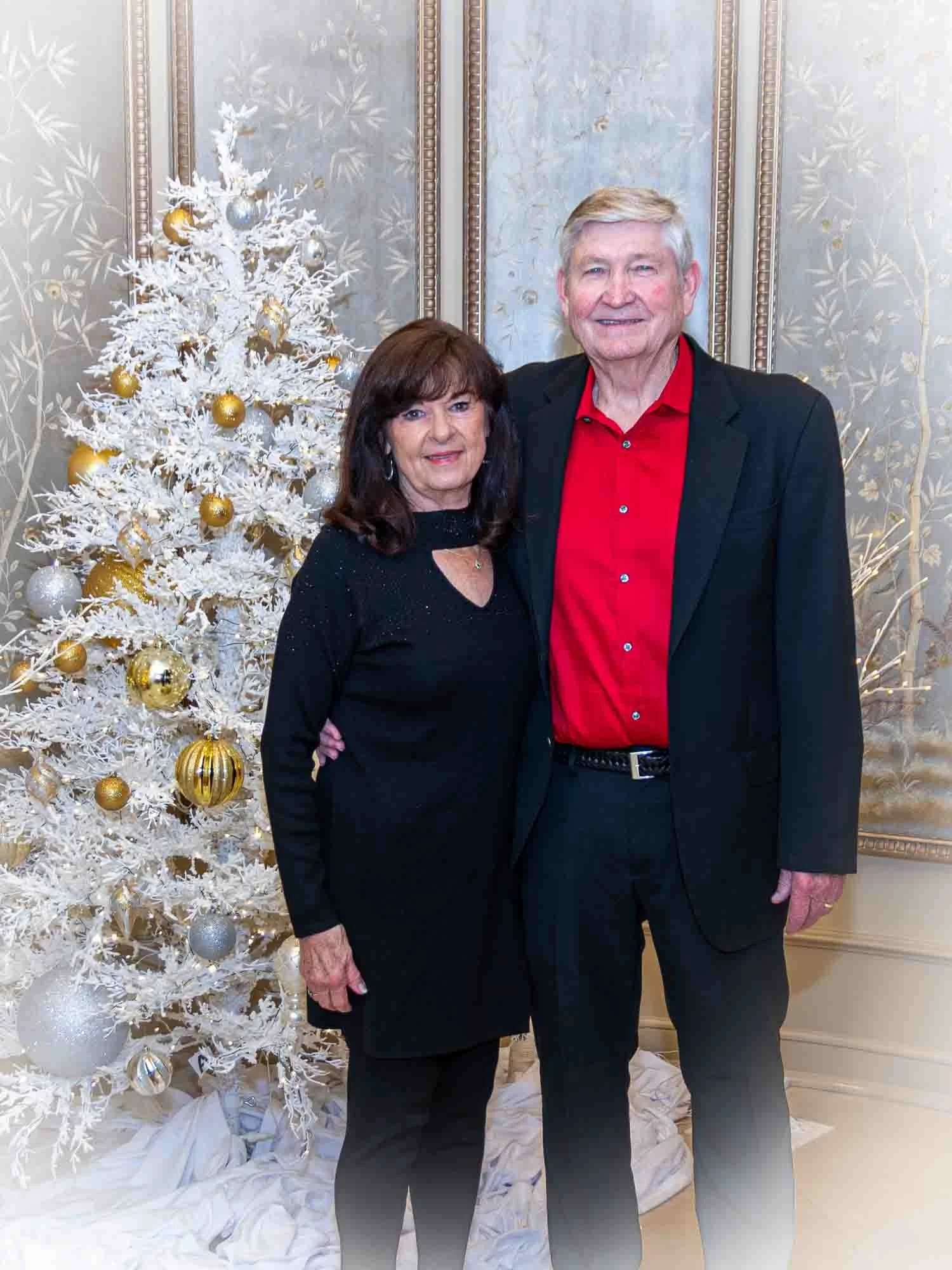 A couple stands smiling in front of a white Christmas tree decorated with gold and silver ornaments. The man is wearing a red shirt and black jacket, while the woman wears a black dress. The setting is elegant with decorative wallpaper and gold-frame
