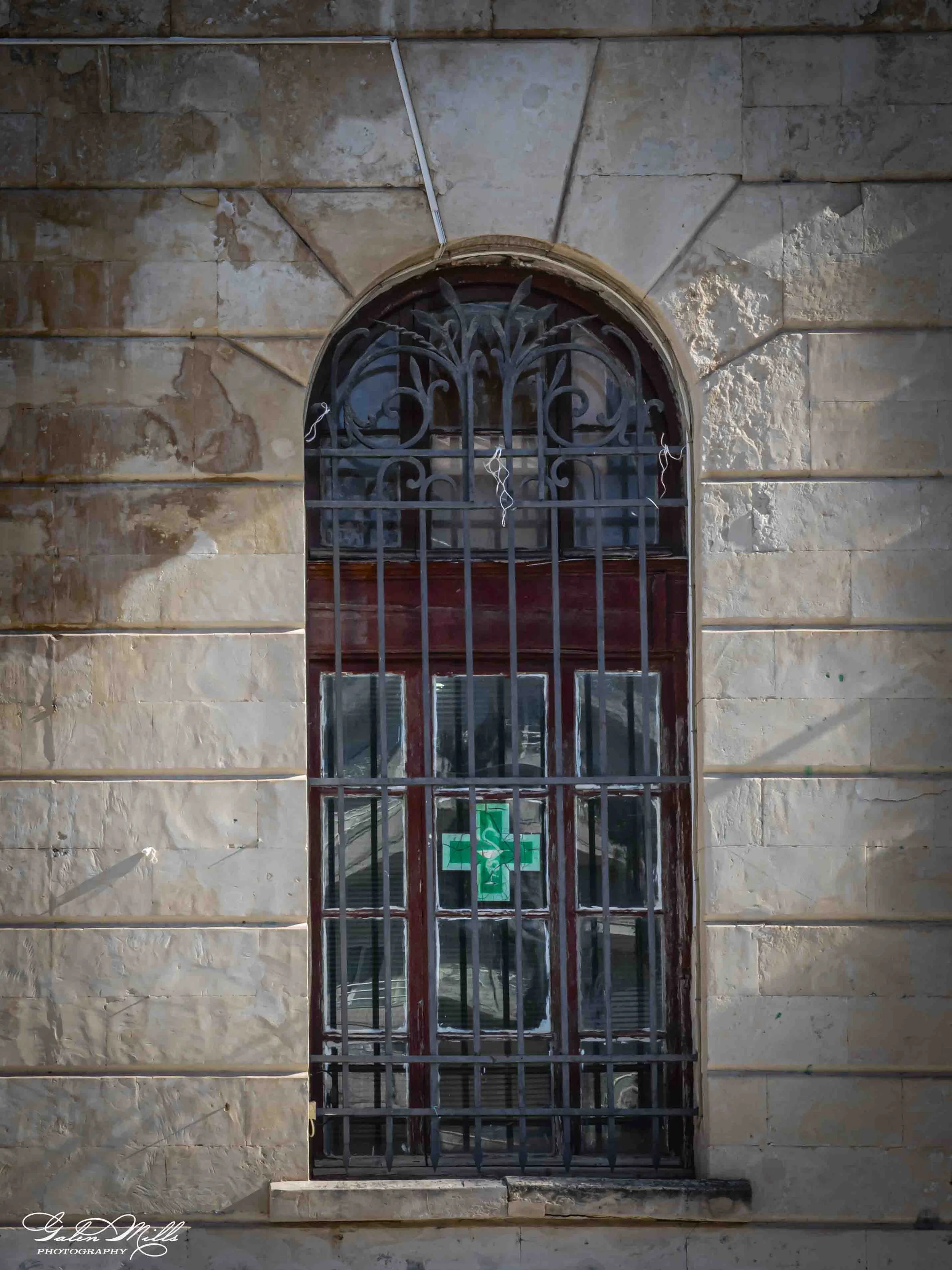 A window with an intricate decorative iron grille on a stone wall with horizontal beige and brown slabs. There is a small ledge beneath the window, and a green cross symbol is visible through the lower window panes.