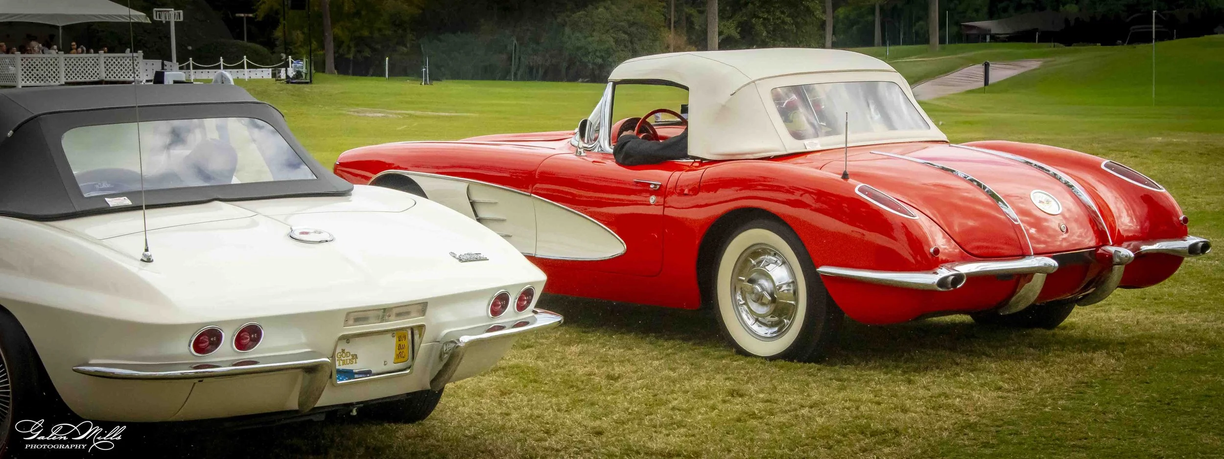 A white vintage convertible car with a black soft top and a red vintage convertible with a white soft top parked on a grassy field, possibly at a car show.
