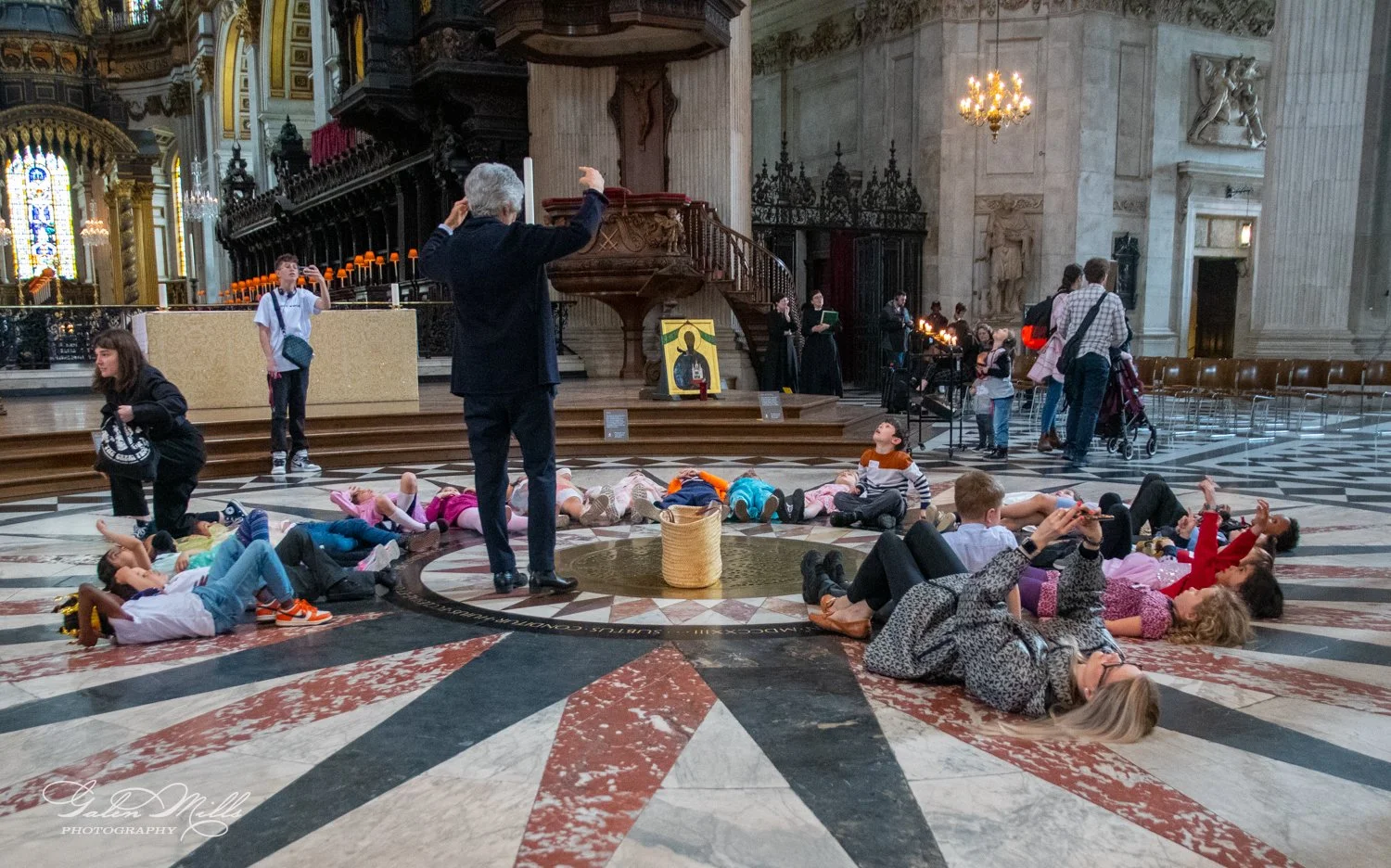 A group of people lies on the floor in a large cathedral, observing the architecture above. An instructor gestures while speaking to them. The surroundings include stained glass windows, a pulpit, and ornate decor.