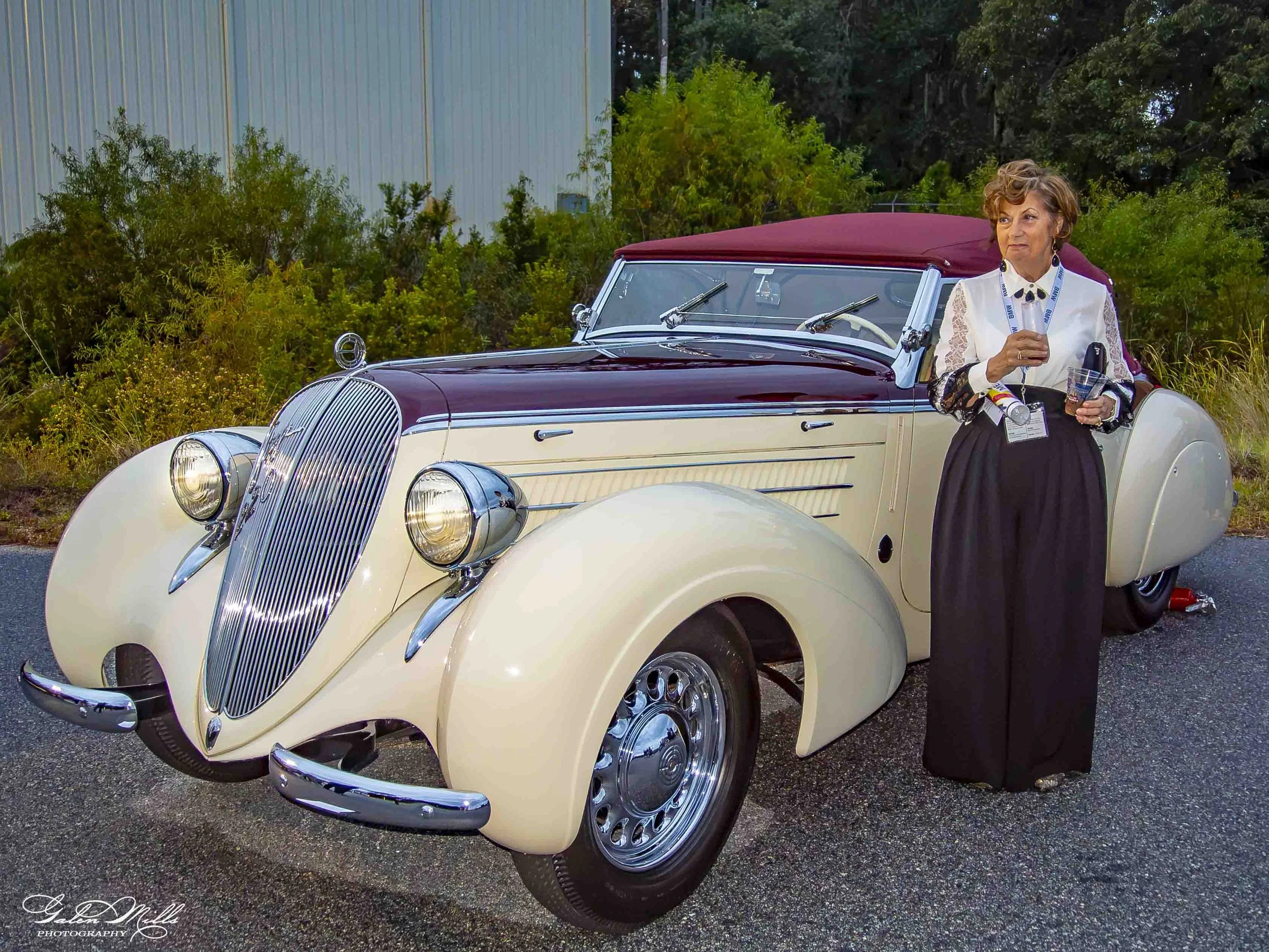 A vintage cream-colored luxury car with chrome details and a maroon convertible top parked outdoors. A woman dressed in formal attire with a lanyard and holding a drink stands beside it, with lush green plants and a blue building in the background.