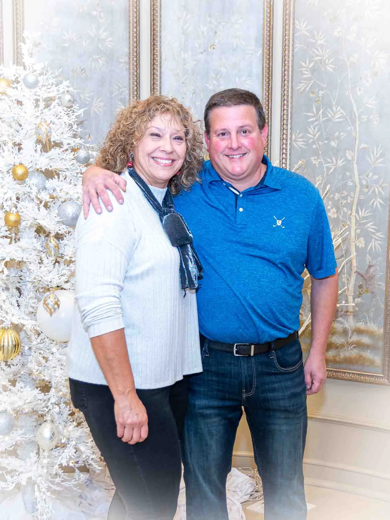 Two smiling people standing beside a decorated white Christmas tree.