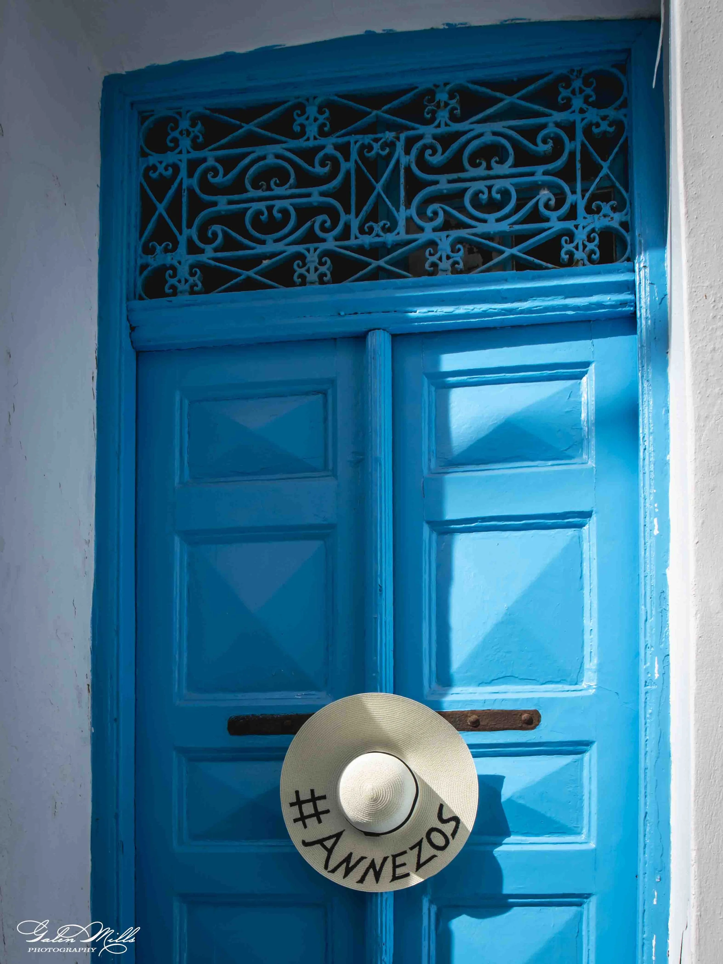 Blue door with decorative ironwork at the top, a white wide-brimmed hat with text '#ALOHA' hanging in front, and a wooden piece at the bottom. The photo has a signature in the lower left corner for Galen Mills Photography.
