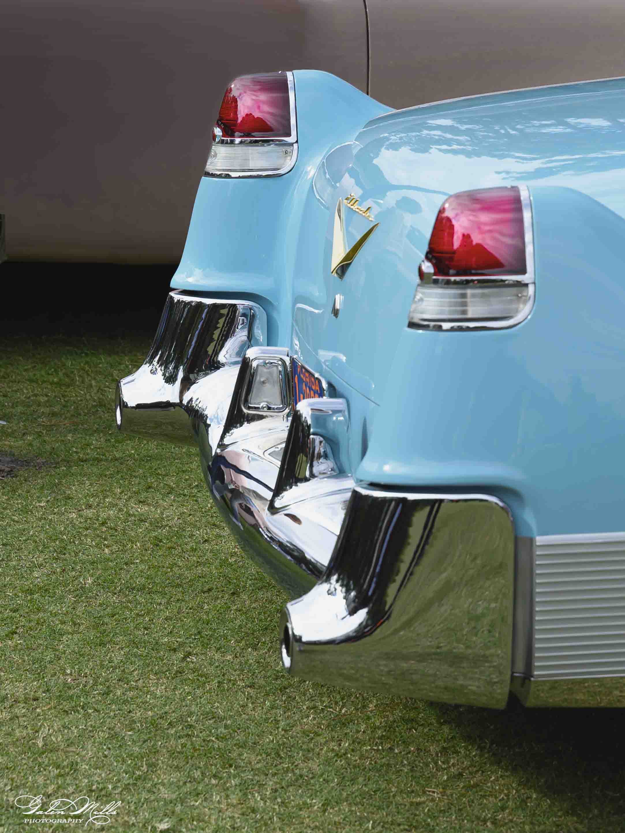 A Cadillac vintage light blue car with chrome accents and tail fins, parked on grass. The back of the car features a red and clear taillight and a gold emblem. The photo is taken outdoors with a neutral background.