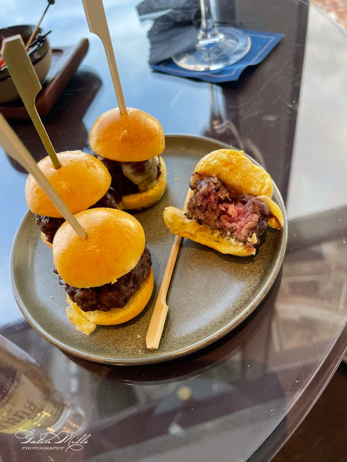 Plate of mini beef sliders with melted cheese and skewers on a glass table.