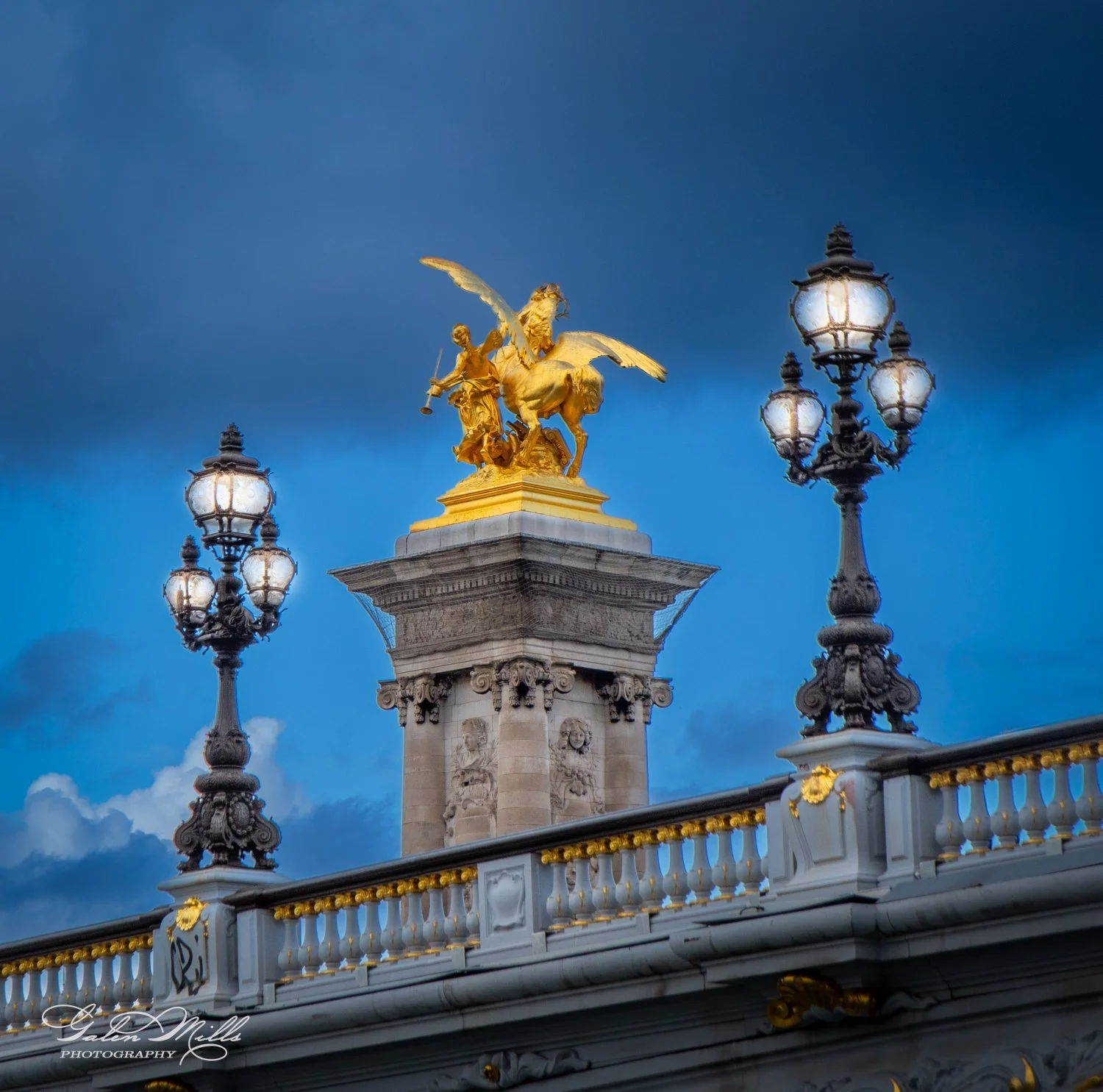 Golden statue of a winged horse on a bridge pillar between ornate street lamps against a dark blue sky.