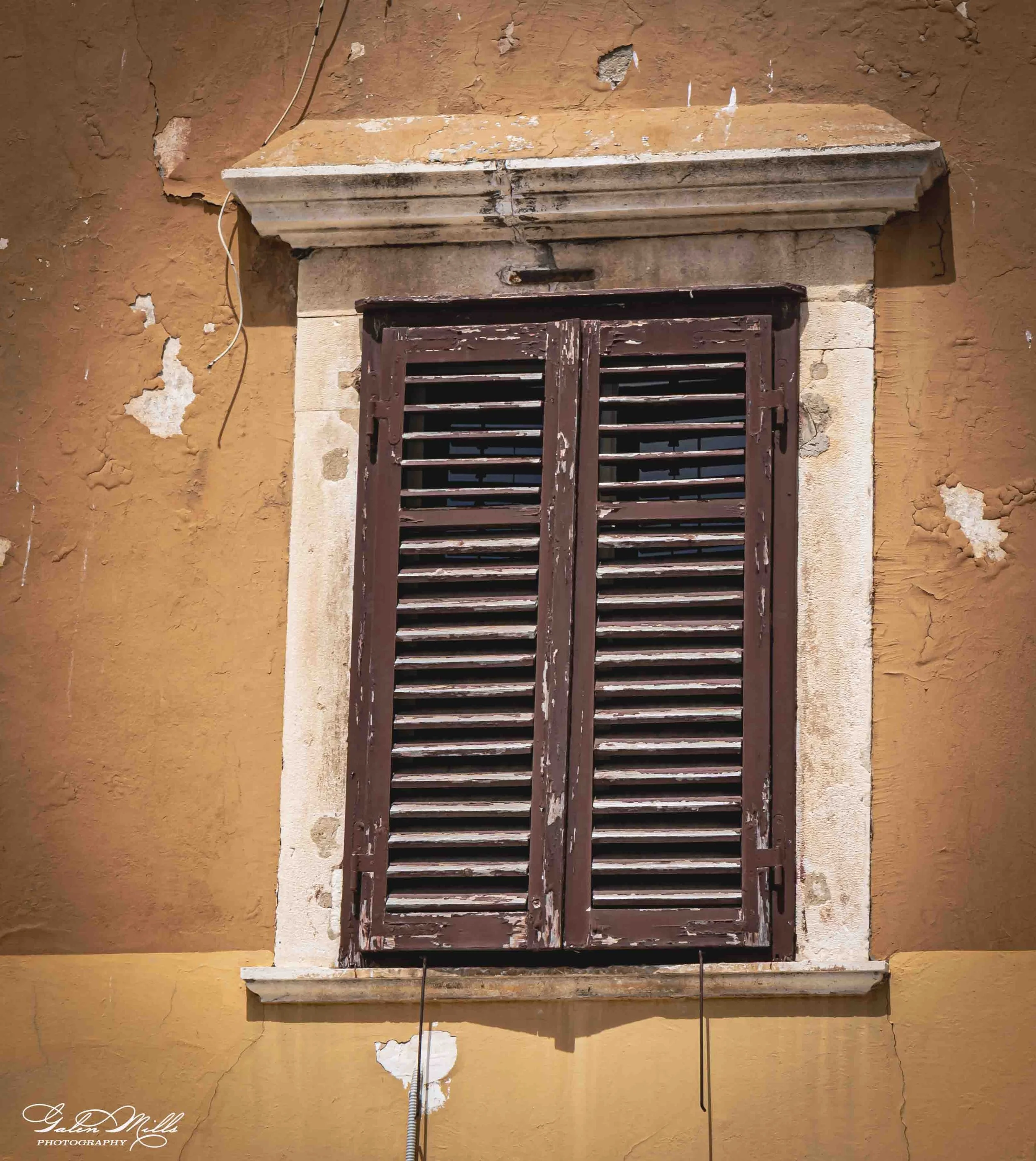 Old, weathered window with closed wooden shutters on a beige, cracked wall with peeling paint.