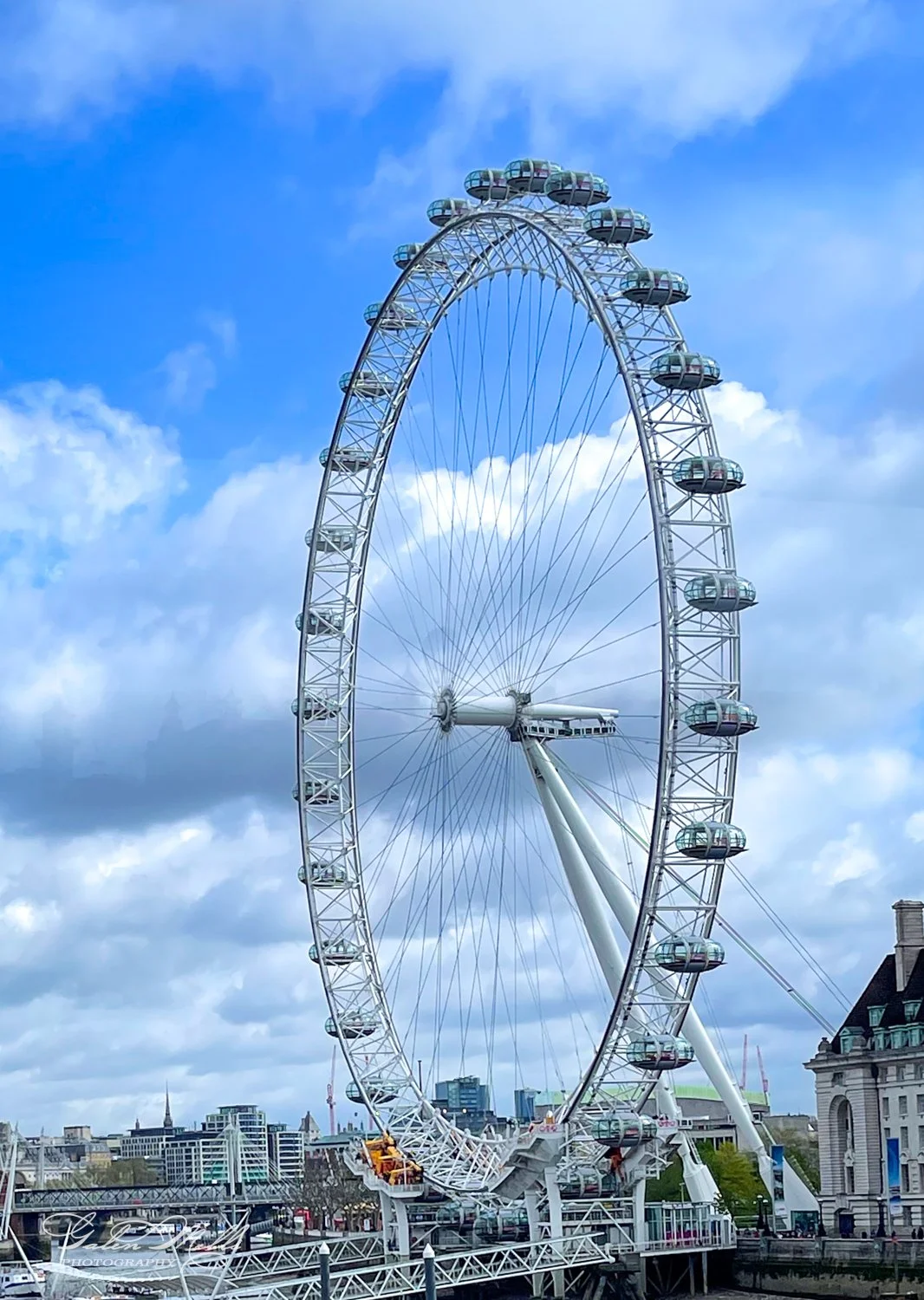 Large Ferris wheel against a blue sky with clouds, cityscape in the background.