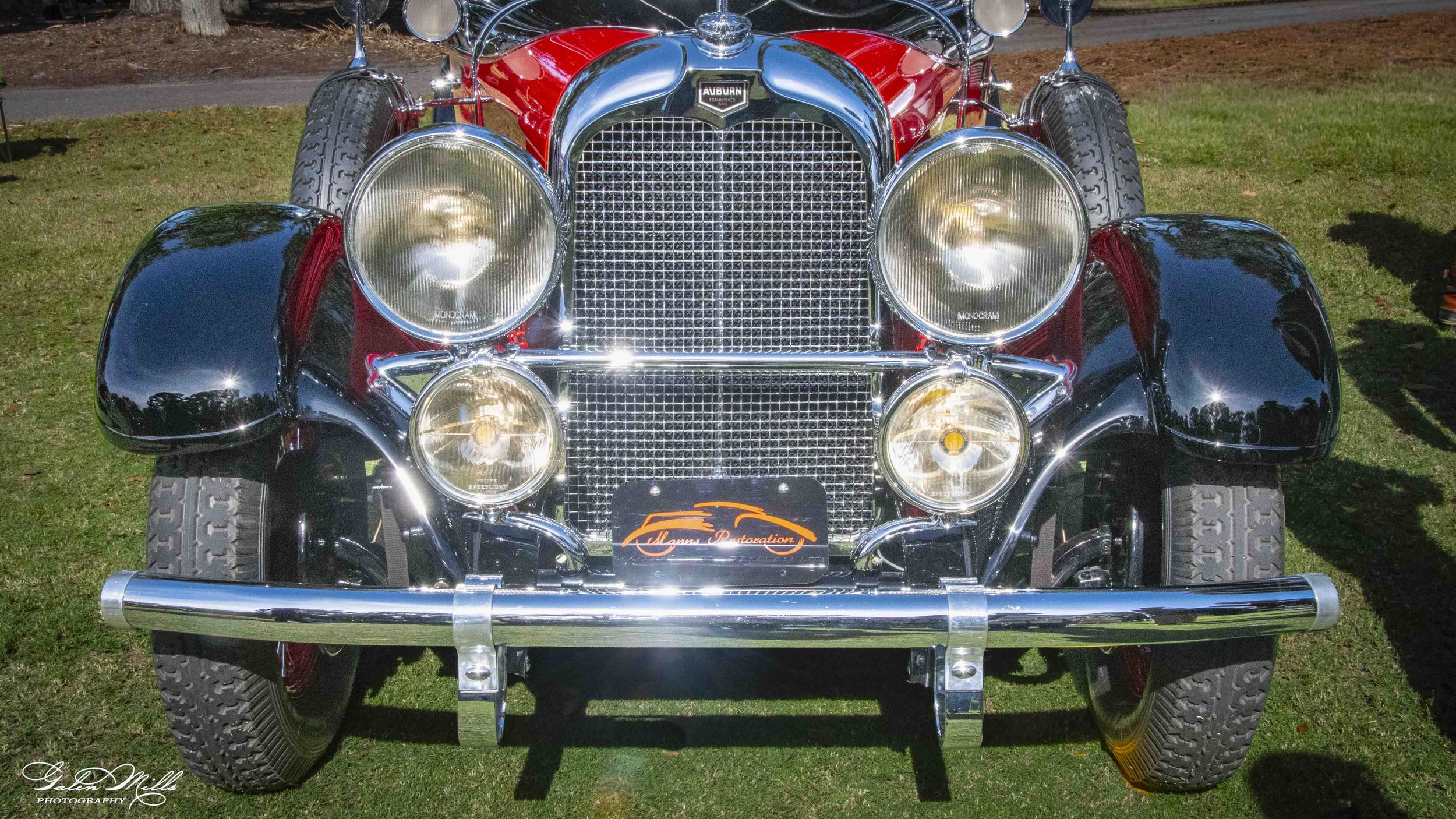 Front view of a vintage red and black car with chrome details and large round headlights, parked on grass.