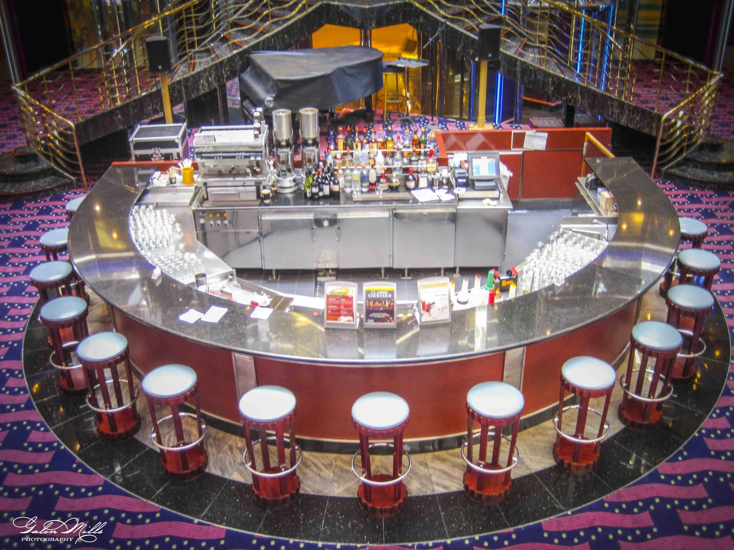 A circular bar area with red and black barstools around a black marble countertop, set on a purple patterned carpet with pink accents, inside a vibrant, upscale venue with curved staircases and colorful lighting.