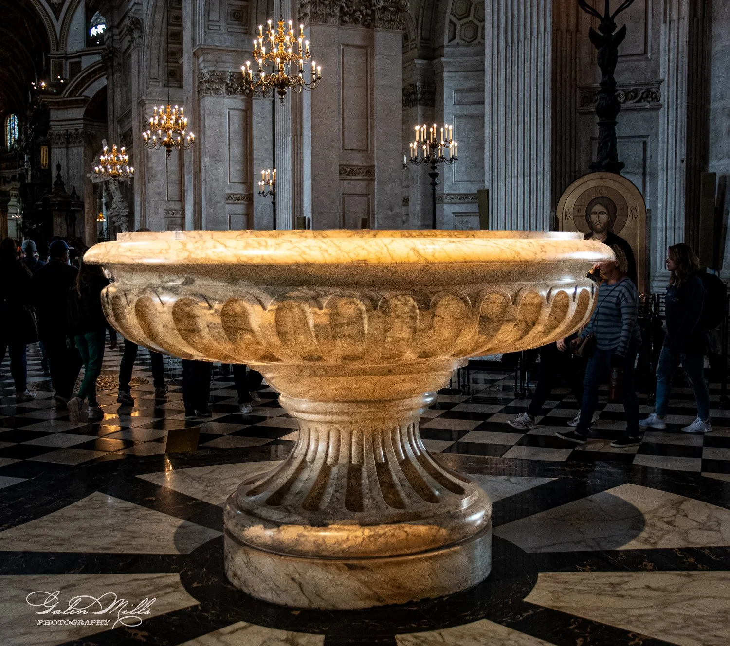 Large ornate marble basin in a grand cathedral interior with chandeliers and people walking around.