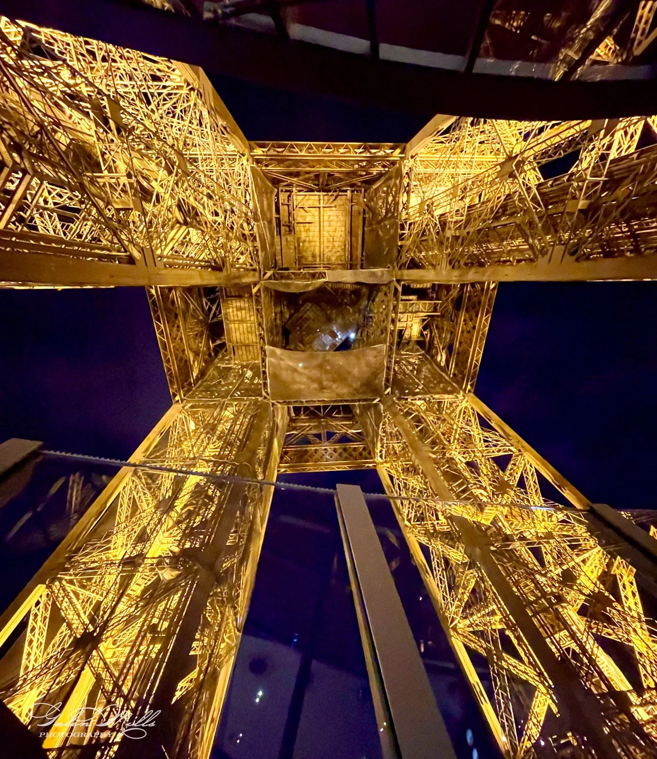View looking up at illuminated Eiffel Tower structure at night, showcasing intricate iron latticework.