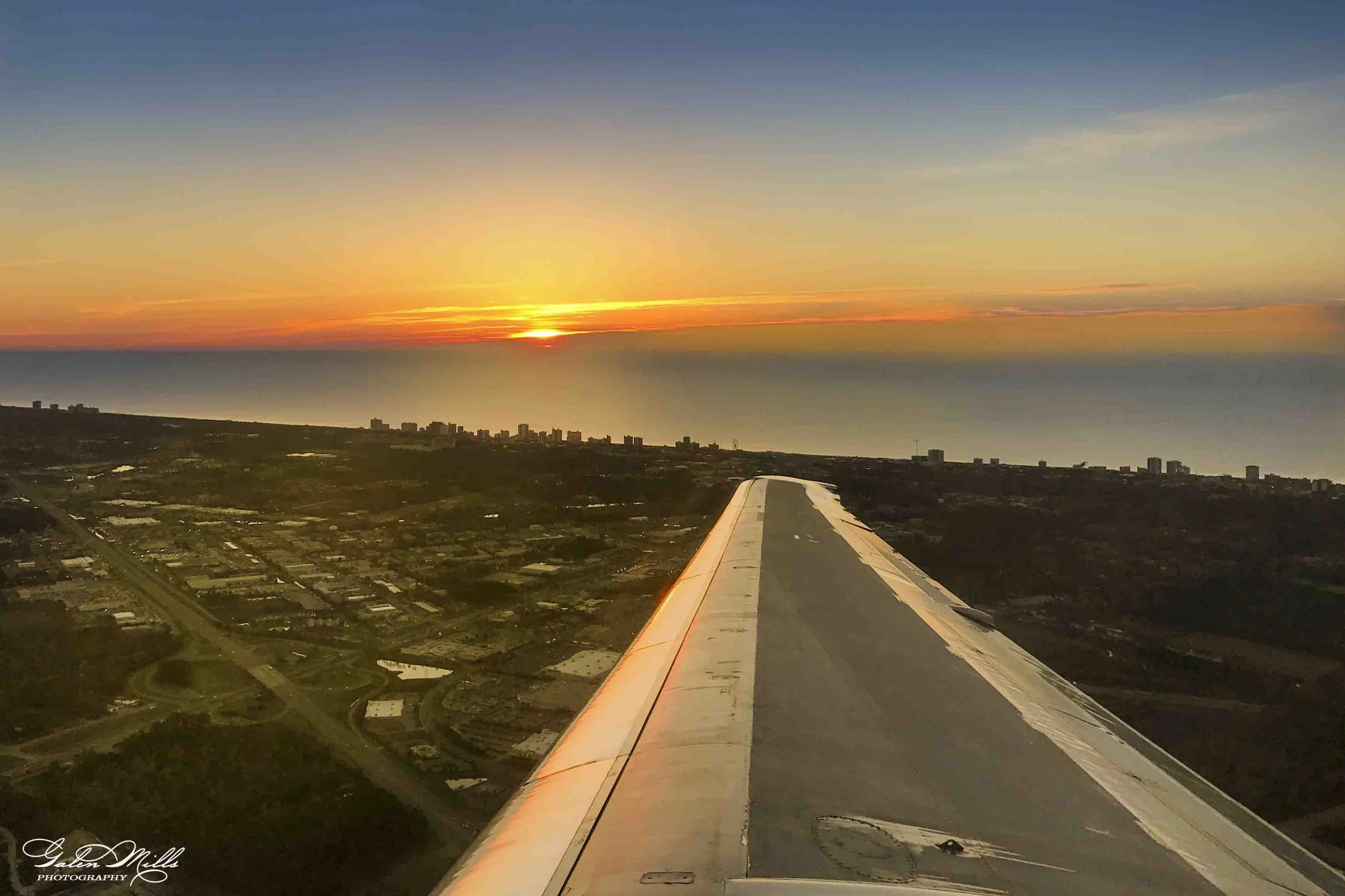 Photo taken from an airplane window showing the wing and a sunset sky over a city with buildings and roads below.