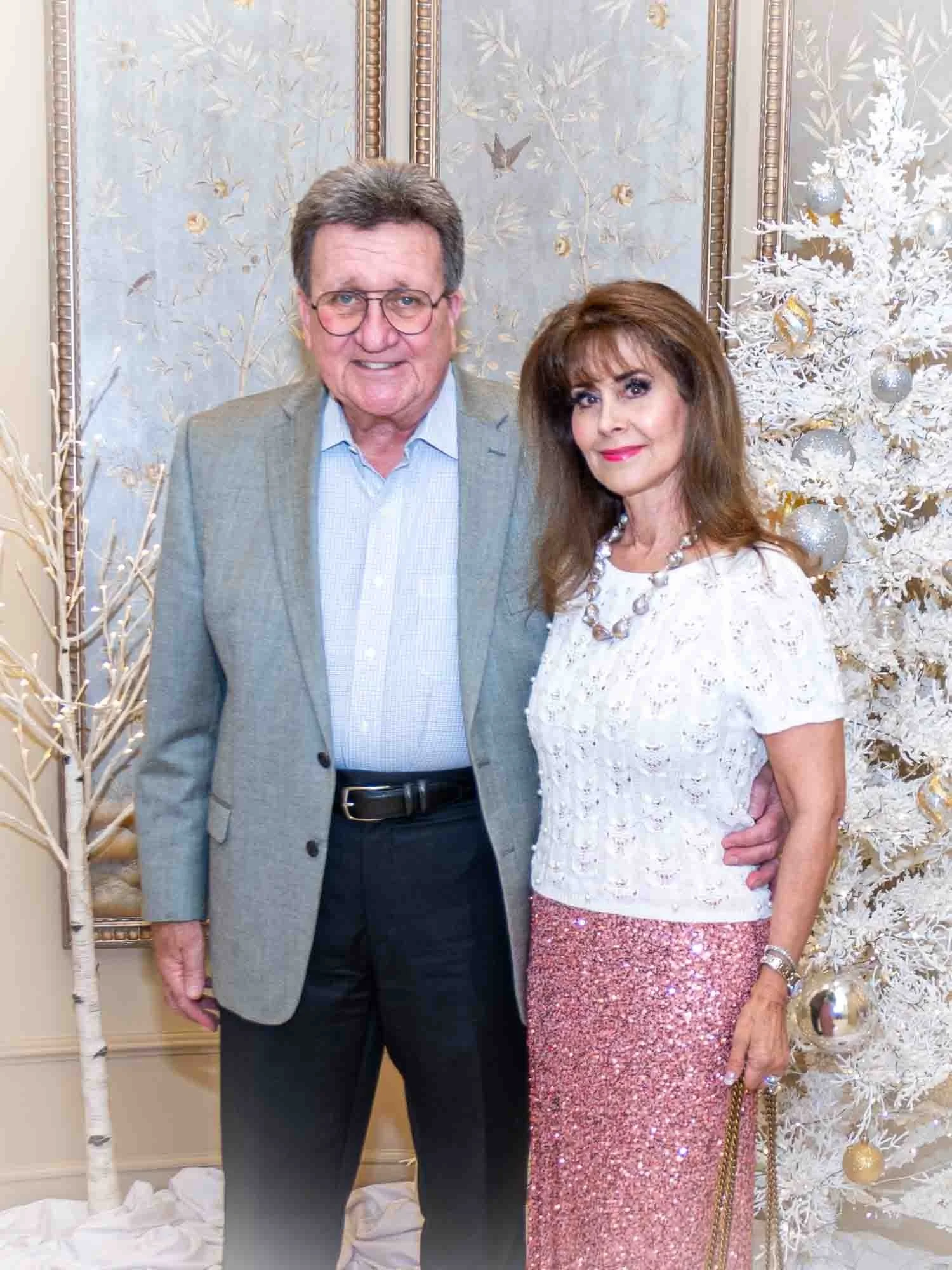 A man in a gray suit and glasses poses with a woman in a white top and pink sequin skirt. They stand in front of a decorative backdrop featuring a white Christmas tree with ornaments.
