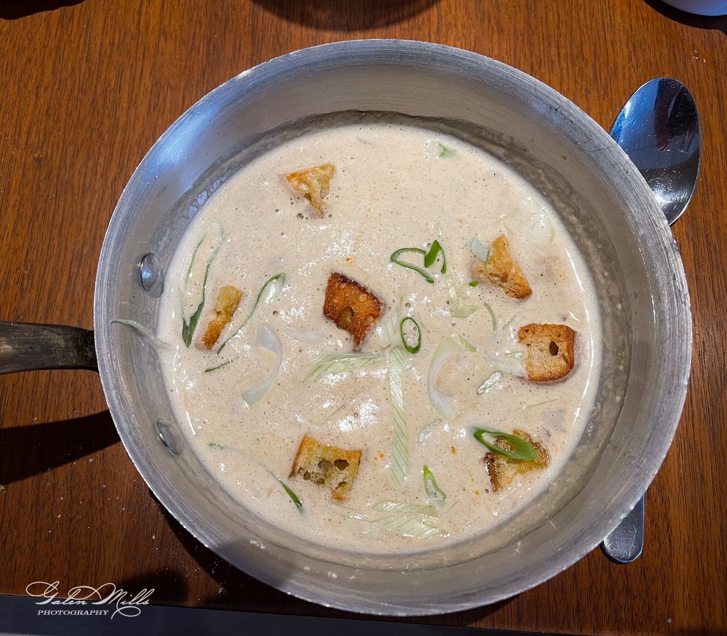 Creamy soup in a pot with croutons and green onions on a wooden table next to a spoon.
