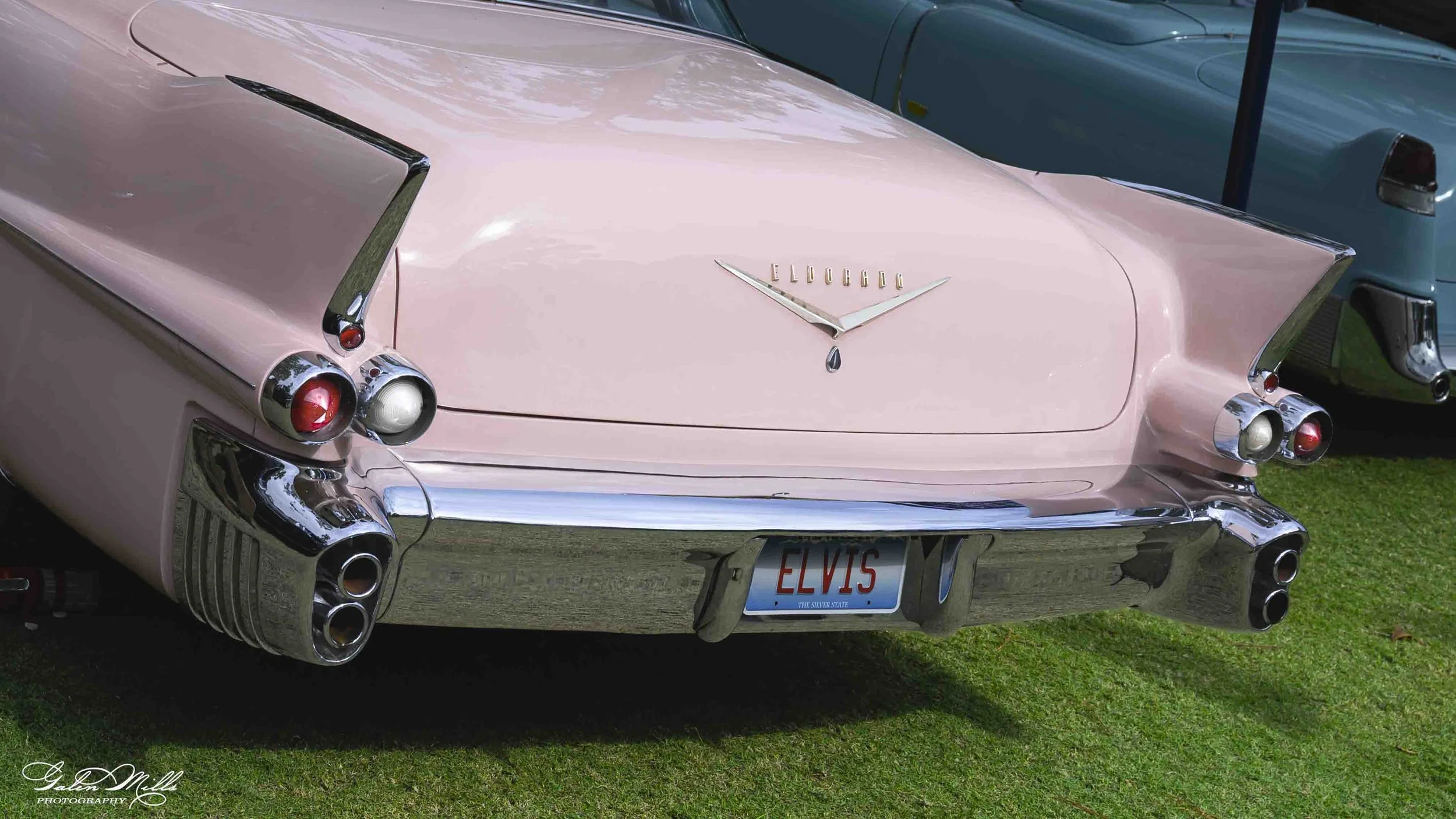 Pink vintage Cadillac car with chrome details and 'Elvis' license plate parked on grass.