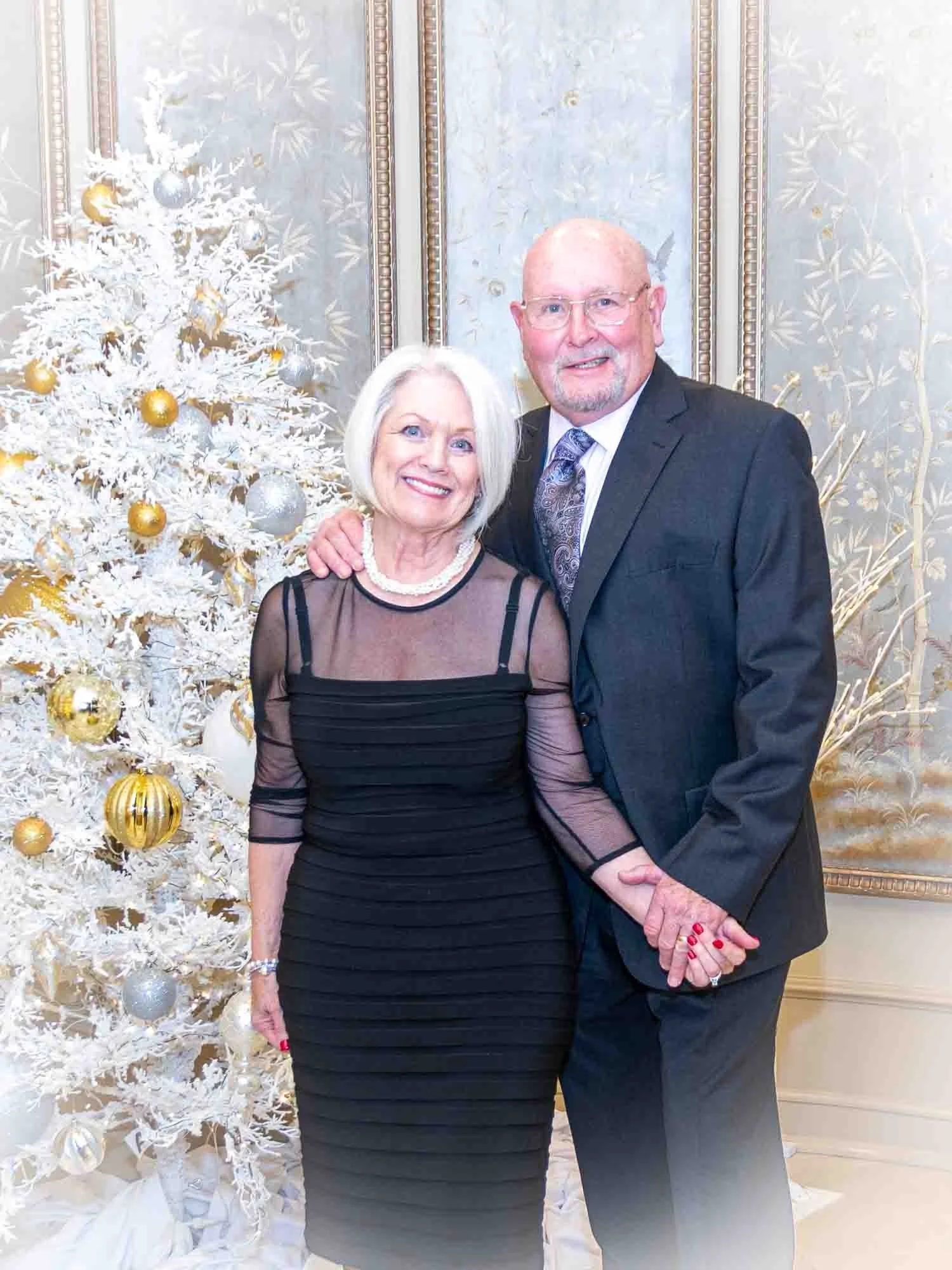 An elderly couple standing together in front of a decorated white Christmas tree with gold and silver ornaments, in a room with ornate wallpaper. The man is wearing a dark suit and tie, while the woman is in a black dress. They are smiling and holdin