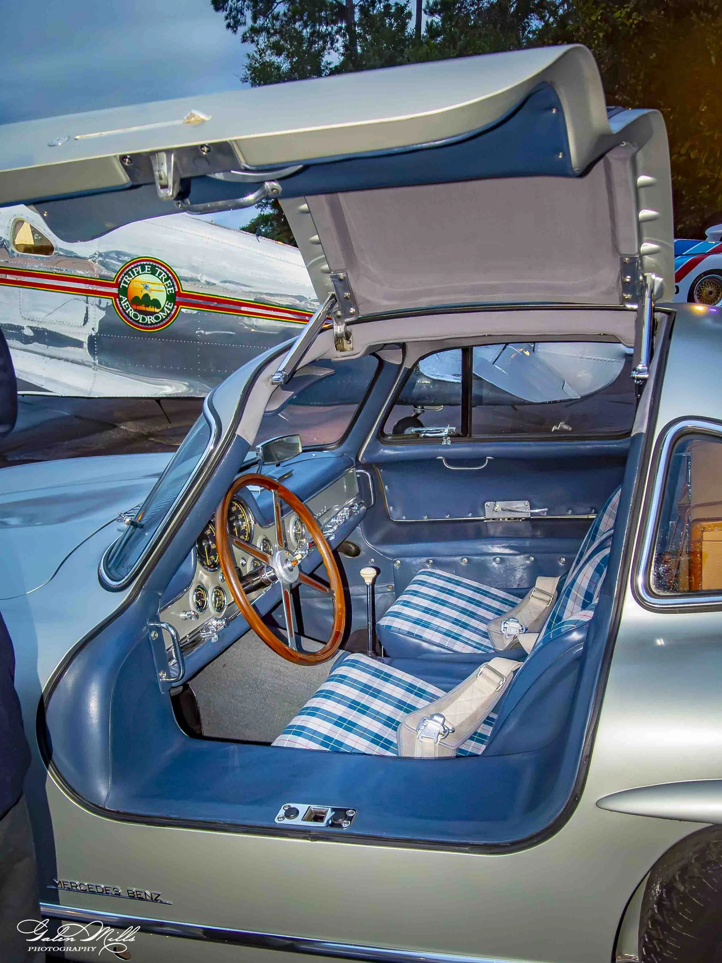 The interior of a vintage Mercedes-Benz car with a blue and grey color scheme, a wooden steering wheel, and plaid patterned seat upholstery, with a rear engine air-cooled plane visible through the open door window.