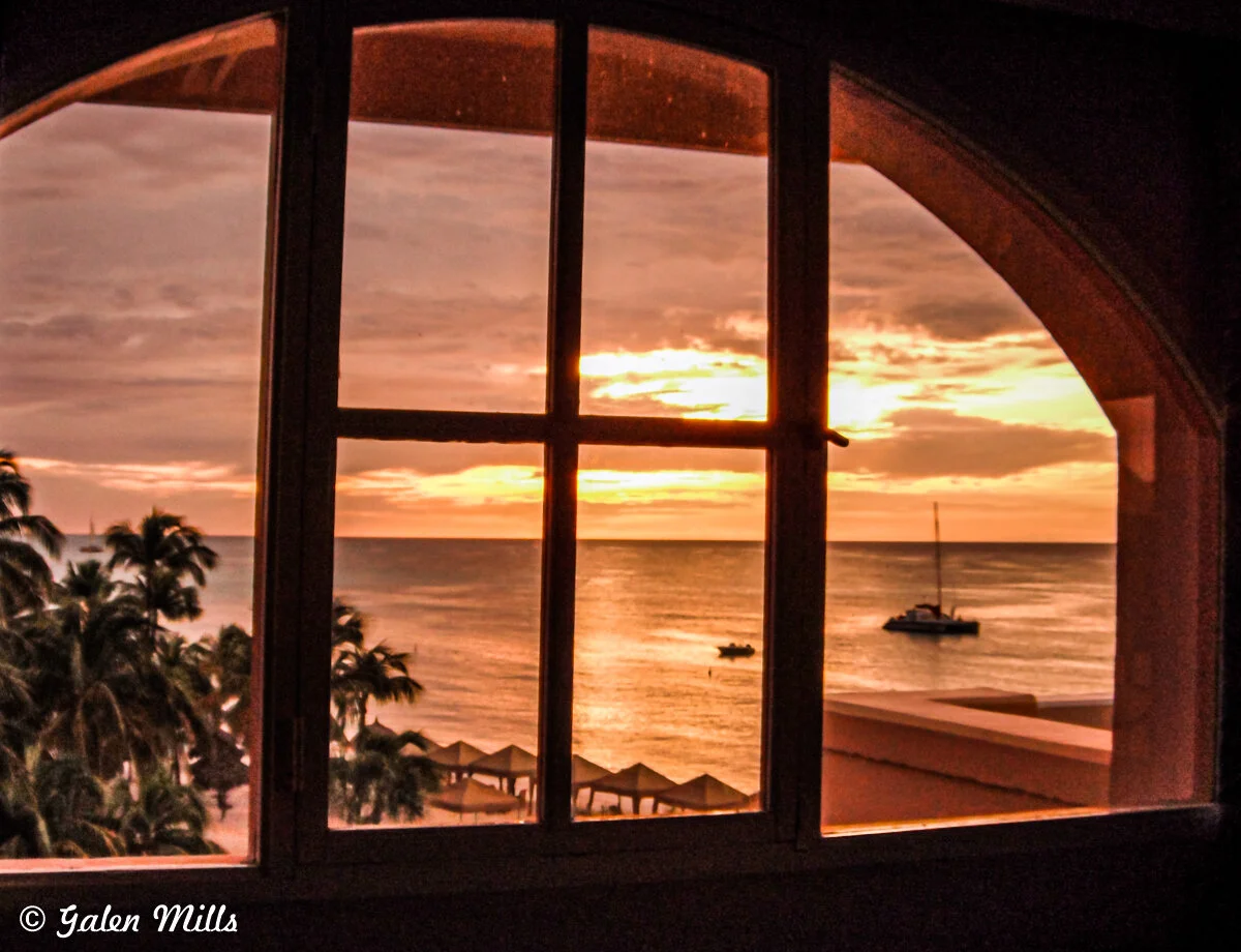 View of a sunset over the ocean through a window, with silhouetted palm trees, beach cabanas, and a sailboat on the water.