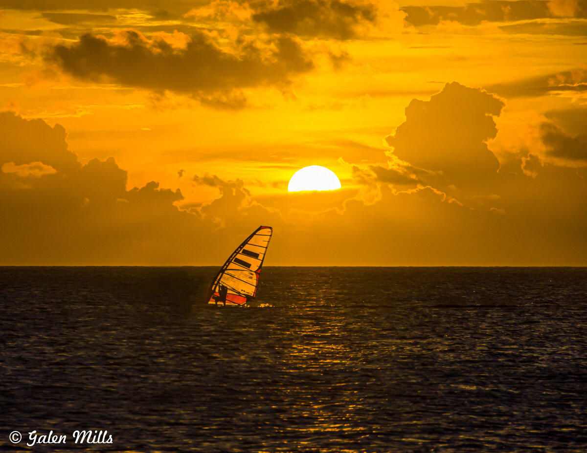 Windsurfer sailing at sunset with orange sky and clouds over ocean.