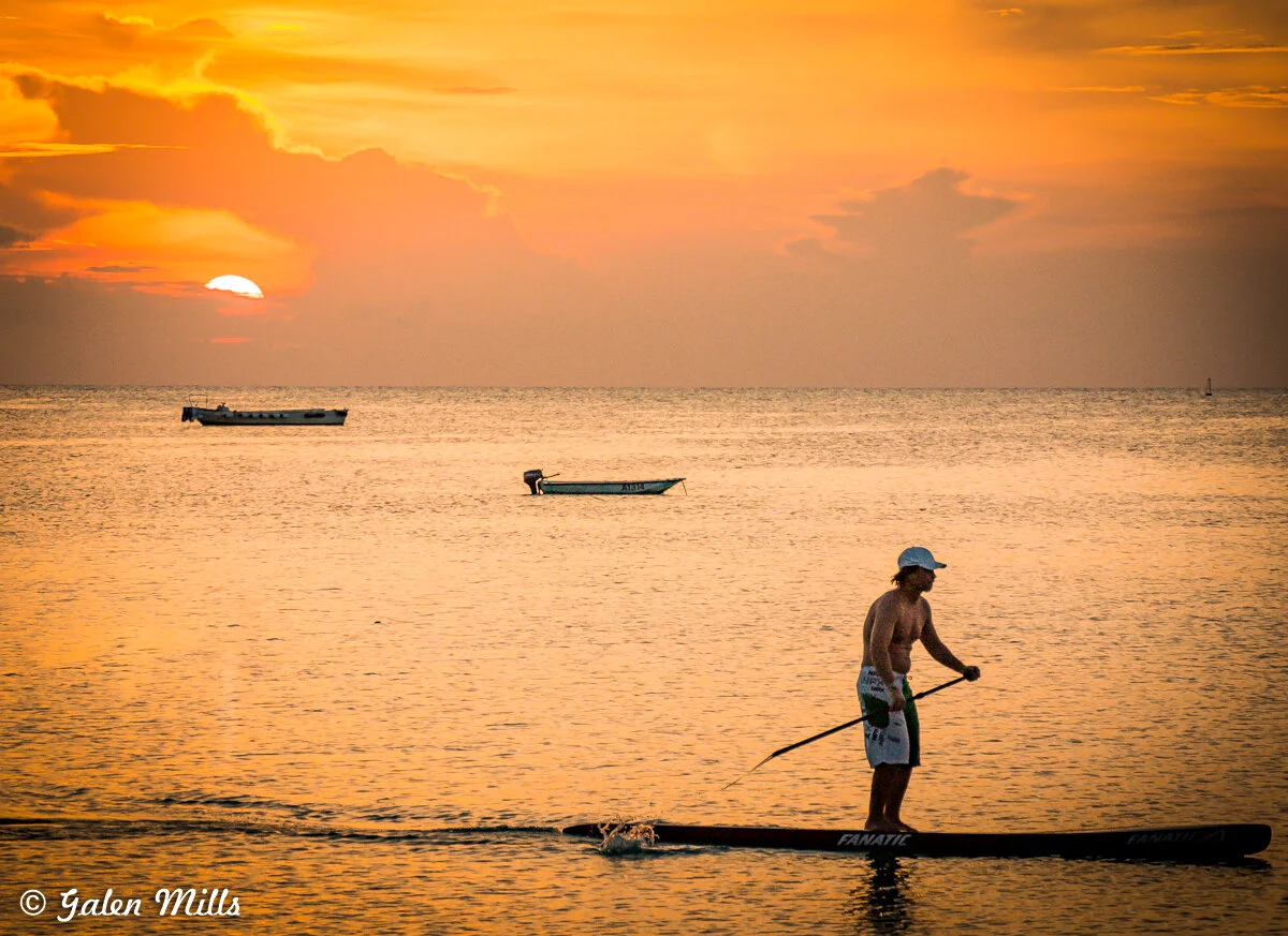 A man paddleboarding on calm ocean waters during a vibrant sunset, with two small boats in the background under an orange sky.