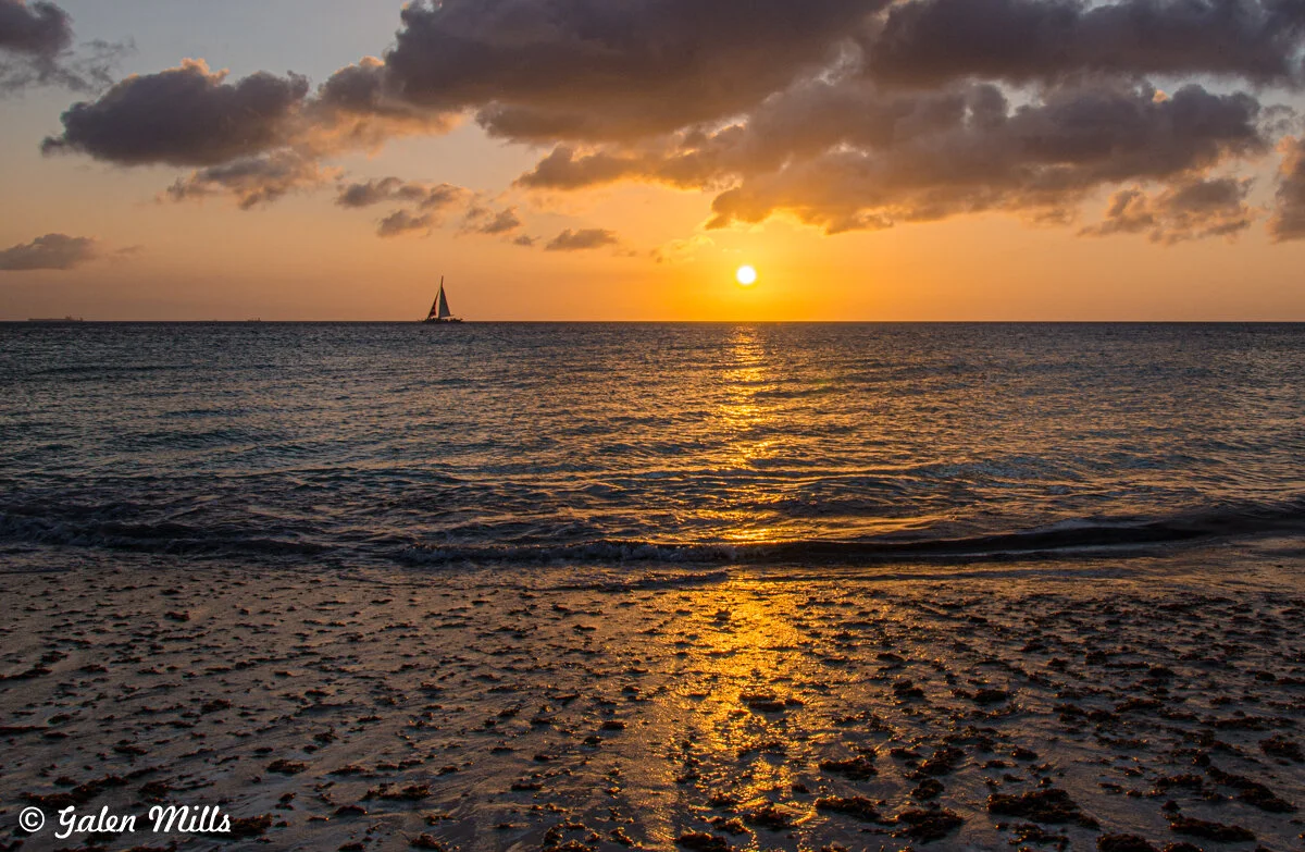 Sunset over the ocean with a sailboat on the horizon, seen from a sandy beach with clouds in the sky.