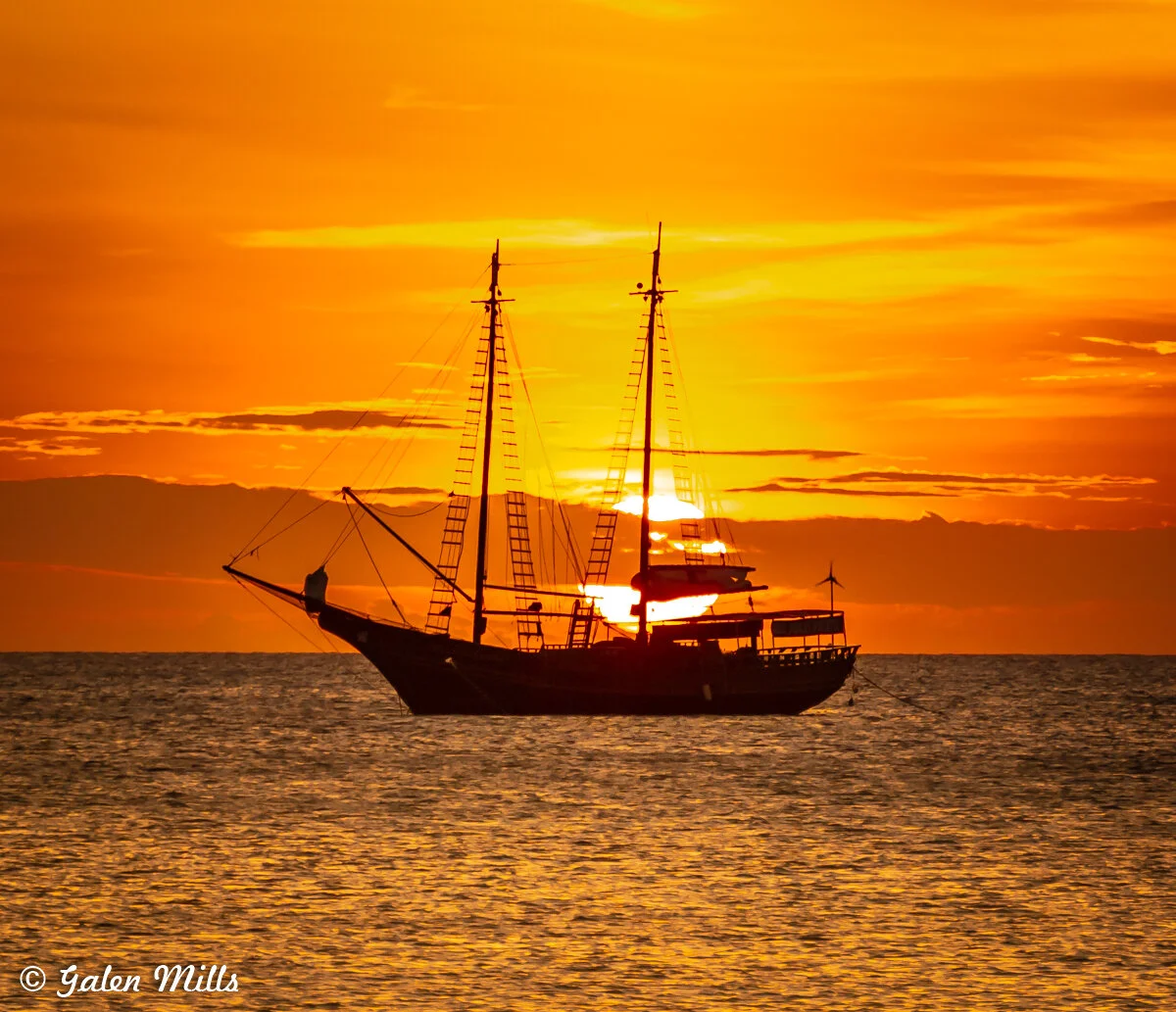 Silhouette of a sailing ship against an orange sunset on the ocean.