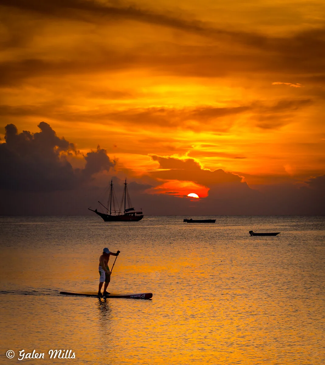 Sunset over a calm ocean with a person paddleboarding in the foreground, silhouetted sailboat and small boats in the background, and a vibrant orange sky with clouds.