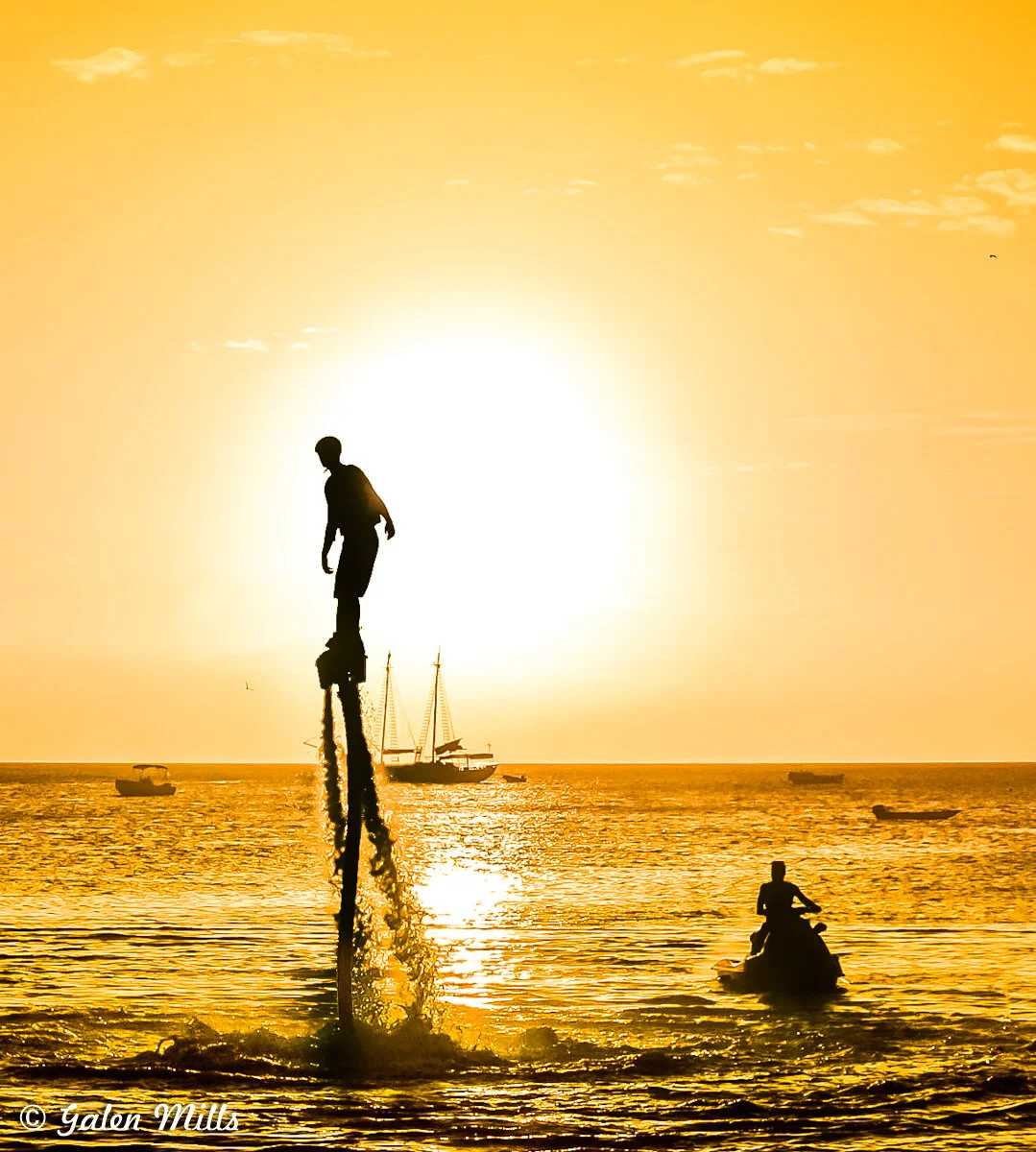 Silhouette of a person flyboarding over the ocean at sunset with a jet ski nearby and boats in the distance.