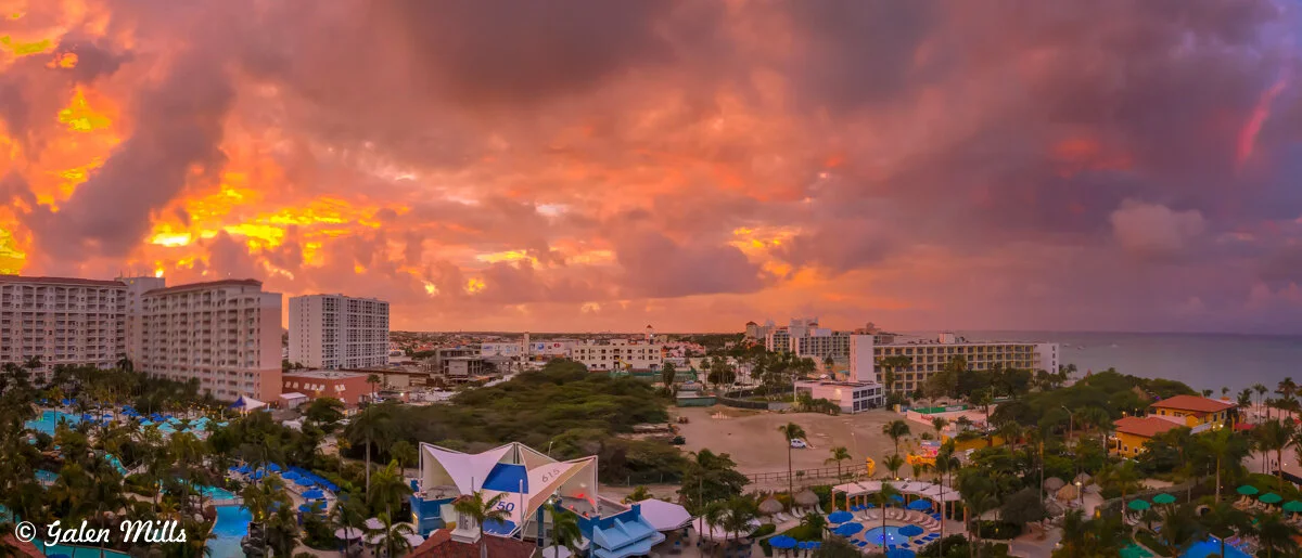 Aerial view of a coastal cityscape at sunset with vibrant orange and pink clouds in the sky. Modern buildings and resorts are surrounded by lush greenery and swimming pools, with the ocean visible in the background.