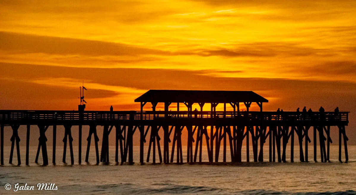 Silhouette of a pier over the ocean at sunrise with orange and yellow skies.