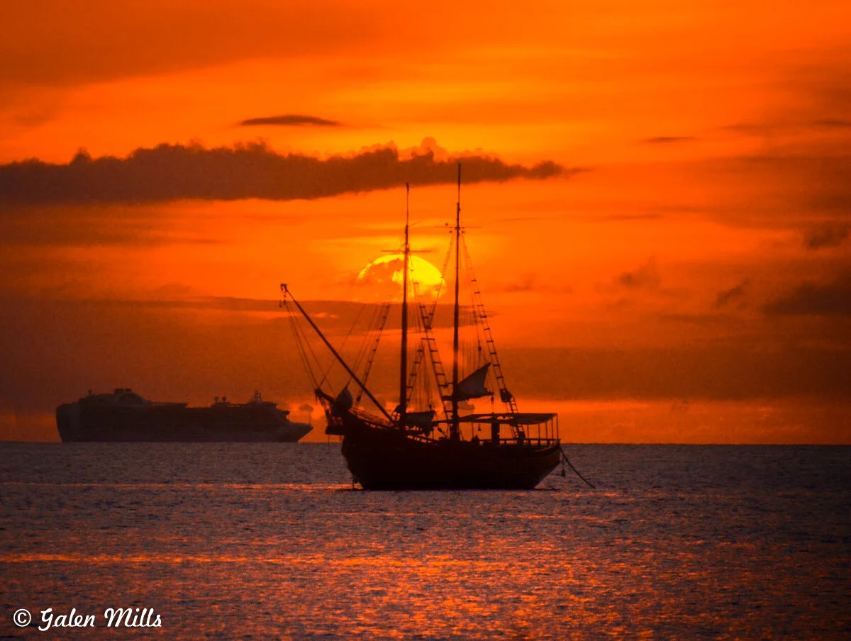 Silhouette of a sailing ship in front of an orange sunset with a cruise ship in the background.