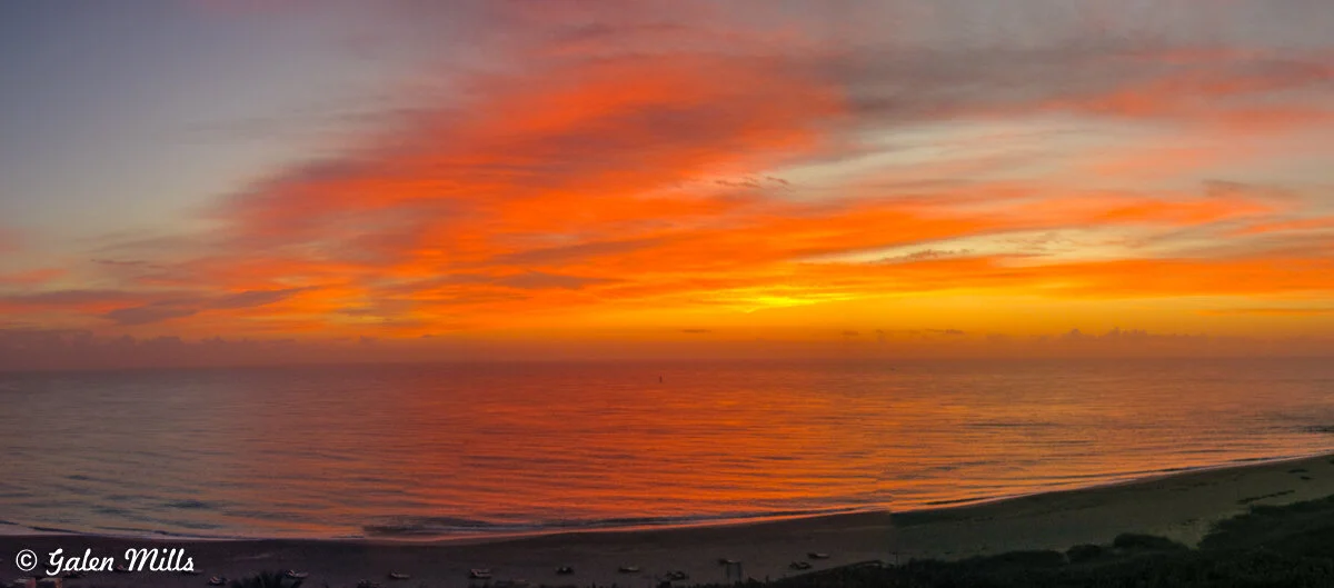 Vibrant sunrise over a calm ocean with an orange and red sky, and a sandy beach in the foreground.
