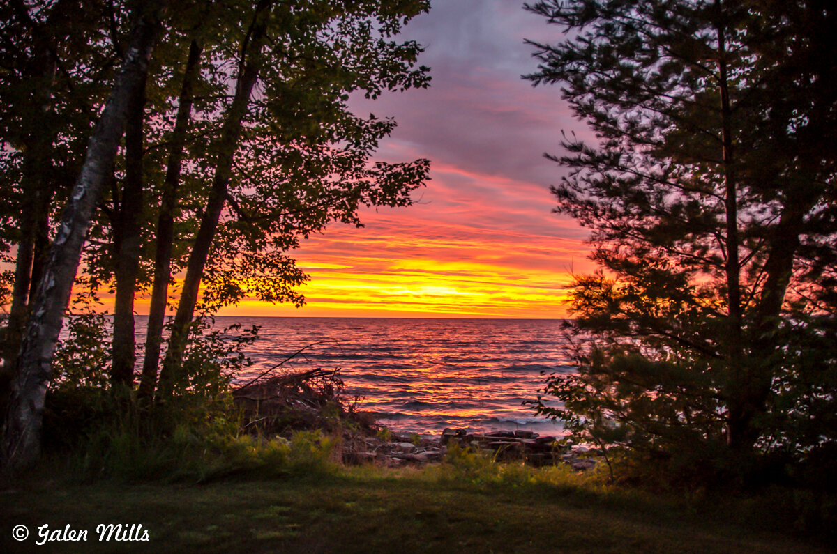 Scenic sunset over a lake, framed by silhouetted trees, with colorful sky reflections on the water.