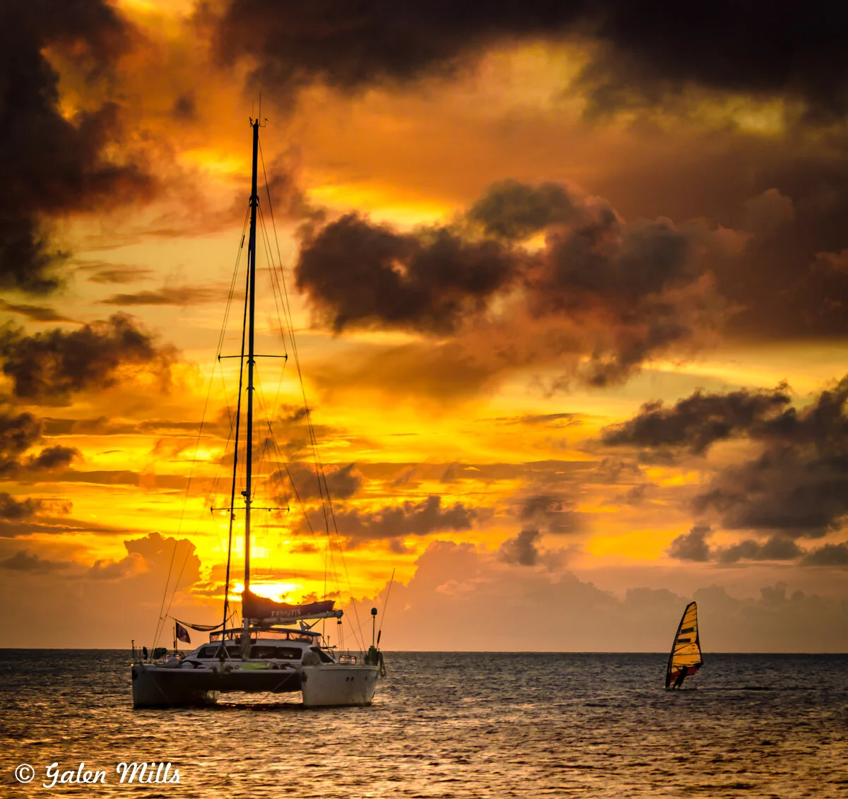 Sailboat and windsurfer on ocean at sunset with colorful sky and clouds.