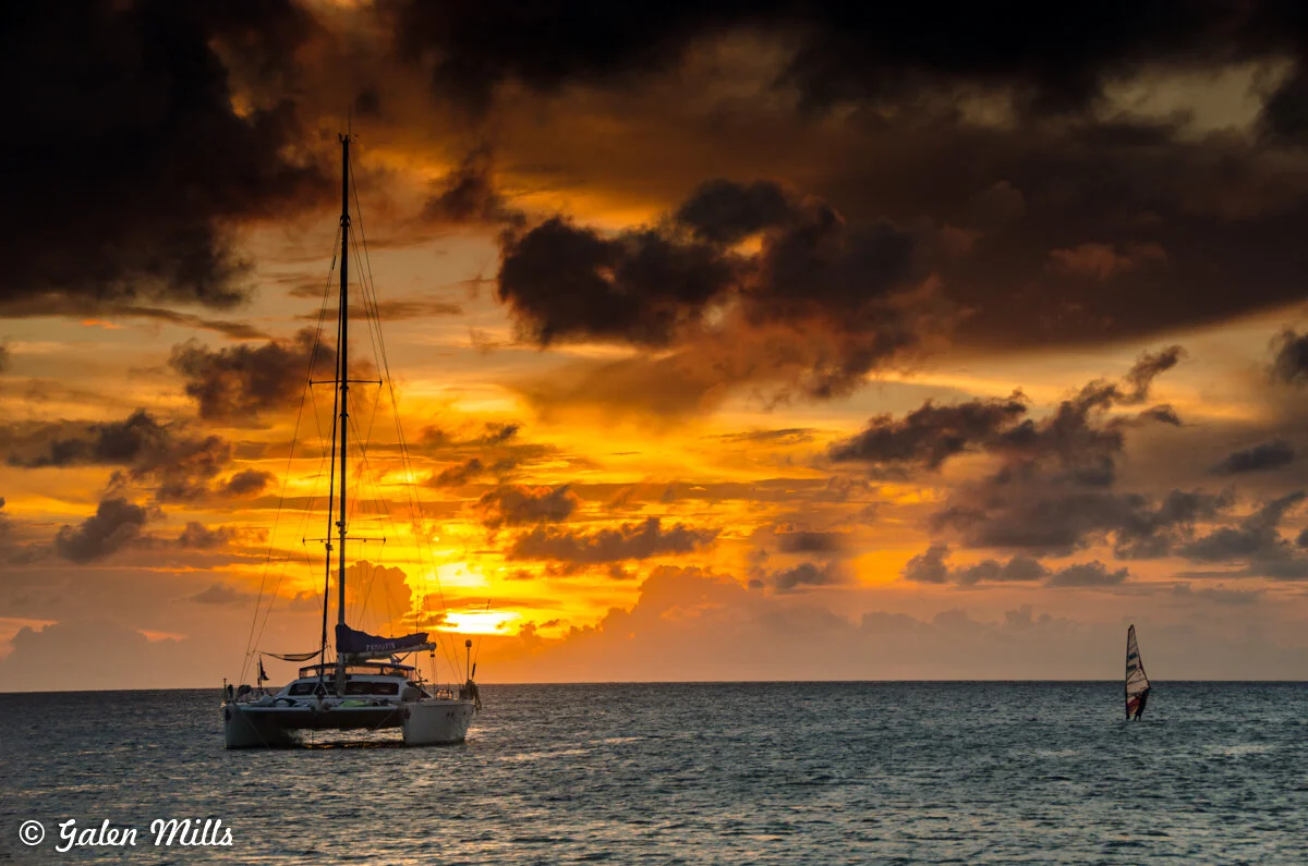 Sailboat and windsurfer on ocean at sunset with dramatic clouds.