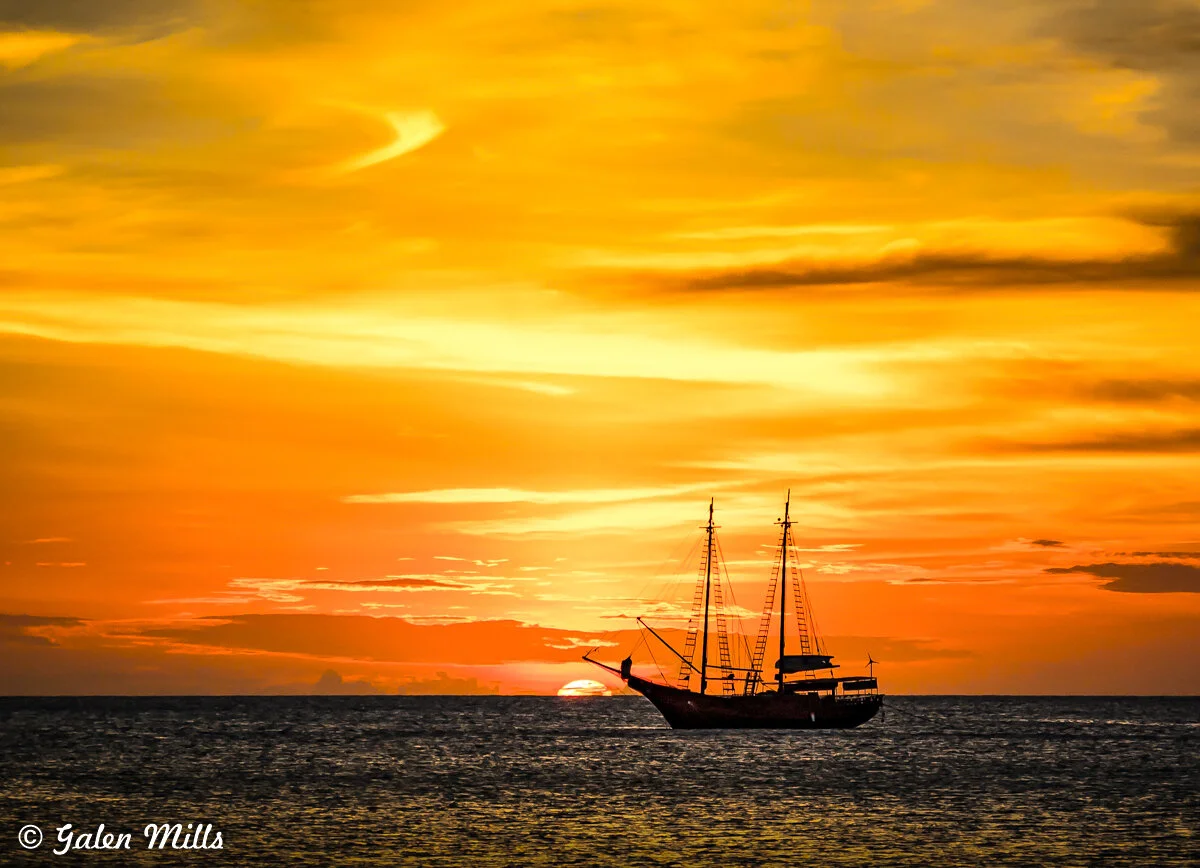 Sailing ship silhouetted against a vibrant orange sunset over the ocean.