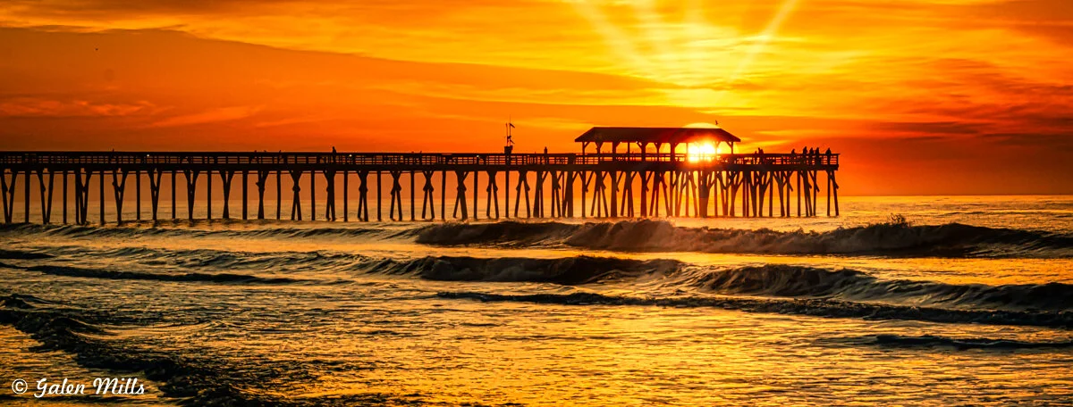 Sunset over an ocean pier with vibrant orange sky and silhouetted structure.
