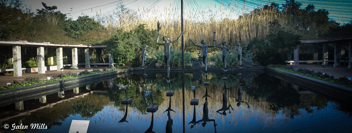 Sculptures of human figures and birds in a reflecting pool surrounded by greenery and columns.